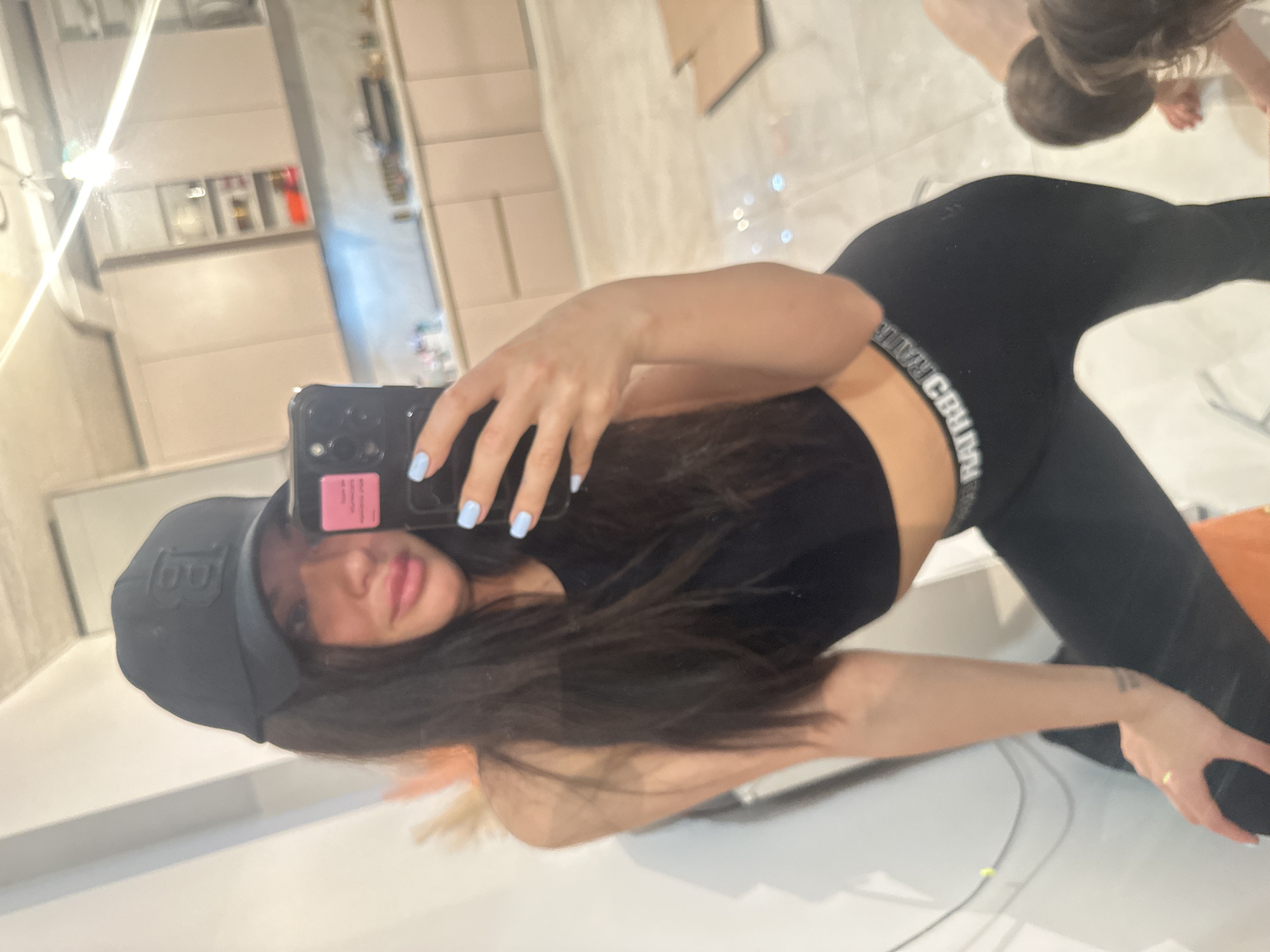A female is taking a mirror selfie in what appears to be an apartment. She is wearing a black baseball cap, a black crop top, and black leggings. Her nails are painted light blue. Another person, possibly a child, is visible in the background reflection. The location is identifiable as the city of Khimki, Russia, based on metadata associated with the image. The kitchen has beige cabinets and is visible in the mirror.