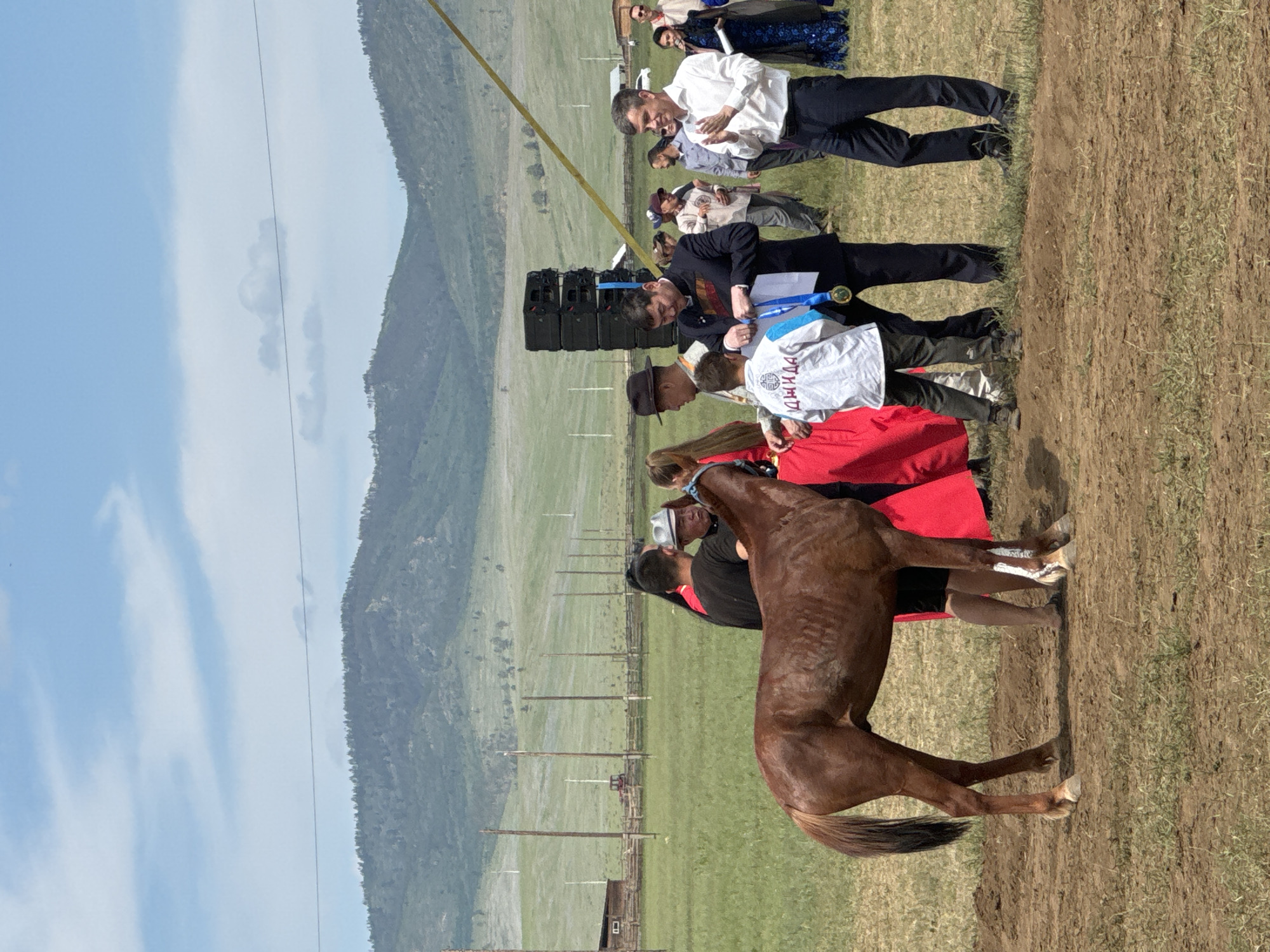 A group of people are gathered around a brown horse in a field with rolling green hills in the background under a partially cloudy blue sky. In the foreground, a man in a white shirt is clapping, facing a man in a black suit who is holding a medal and ribbon. A young boy in a white shirt with blue trim is standing between them, looking at the medal. To the left, a man in a black shirt and a woman in a red dress are standing near the horse. Further back, a stack of black speakers is visible, as well as tents and fencing. The overall scene appears to be an outdoor award ceremony or event, possibly in Orongoi, Russia, given the mountainous terrain and style of dress.