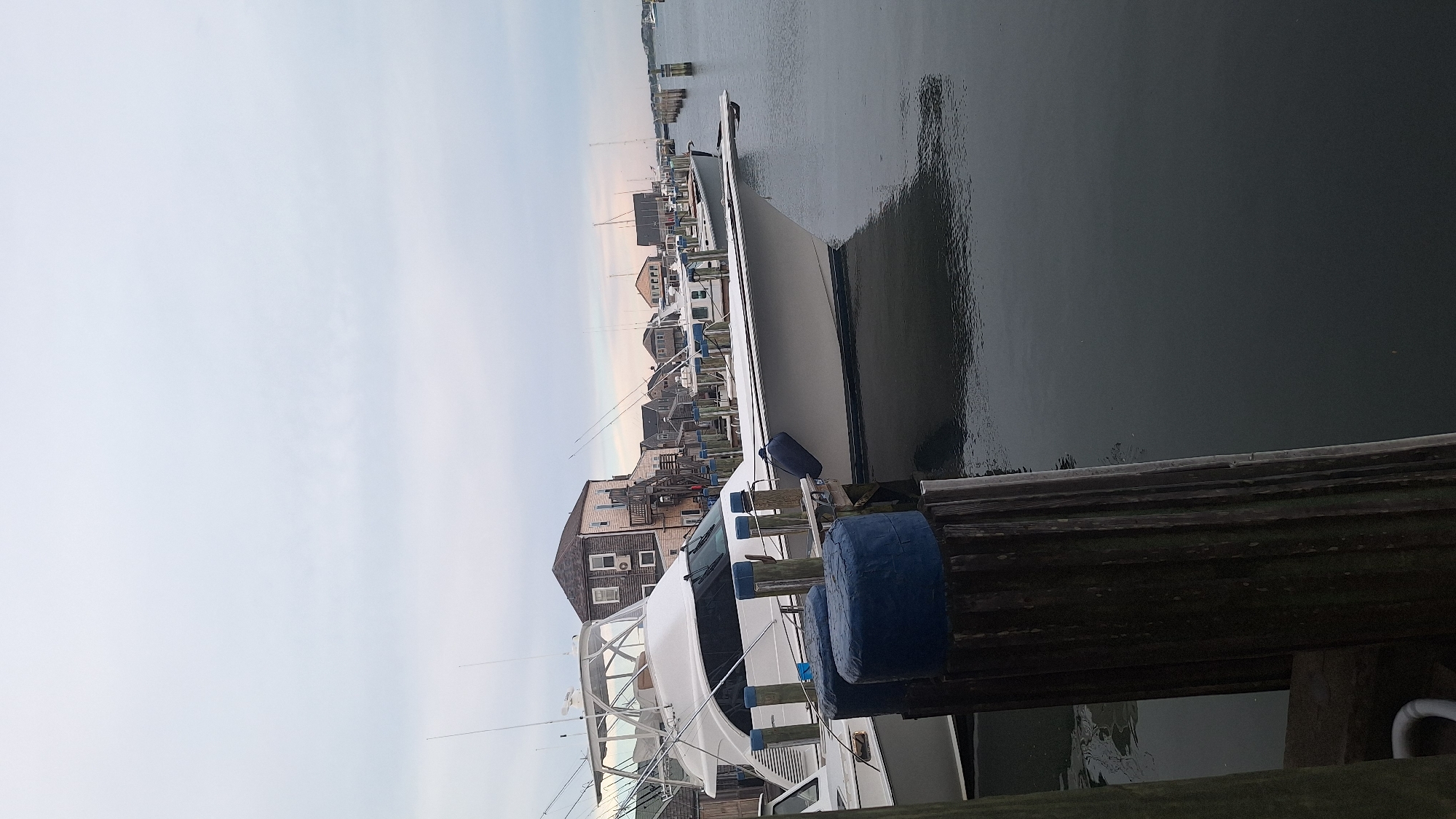 The image shows a waterfront scene with several boats docked at a pier in Nantucket, United States of America. The sky is overcast, and the water is calm. In the foreground, there are wooden pilings with blue protective coverings. A large white yacht with a hardtop is prominently featured, docked alongside other vessels. Behind the boats, a row of buildings with traditional New England architecture is visible. The overall atmosphere is tranquil and suggests a quiet harbor setting. There are no people visible in the image.