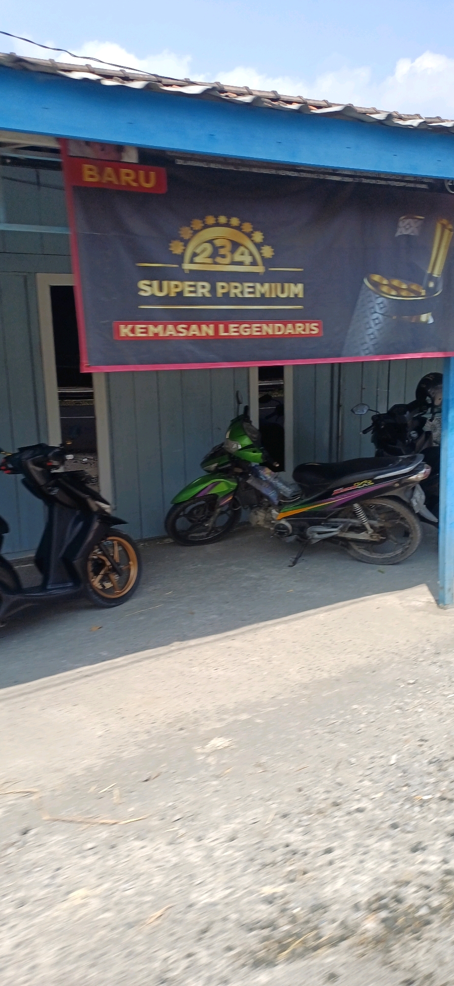 The image depicts the exterior of a shop in Jakenan, Indonesia, under a bright sky with scattered clouds. A dark banner with gold lettering and graphics is prominently displayed across the front of the building. The banner reads "BARU" in red on the left side, followed by "234 SUPER PREMIUM KEMASAN LEGENDARIS" in gold on a dark background. To the right of the text, there is an illustration of a golden chalice or goblet.
In front of the shop, two motorcycles are parked on a paved surface. The motorcycle on the left is black with gold wheels. The motorcycle on the right is green and black. The building itself appears to be made of light blue wooden planks. The overall scene suggests a retail establishment, likely a convenience store or a small business, in a local Indonesian setting.