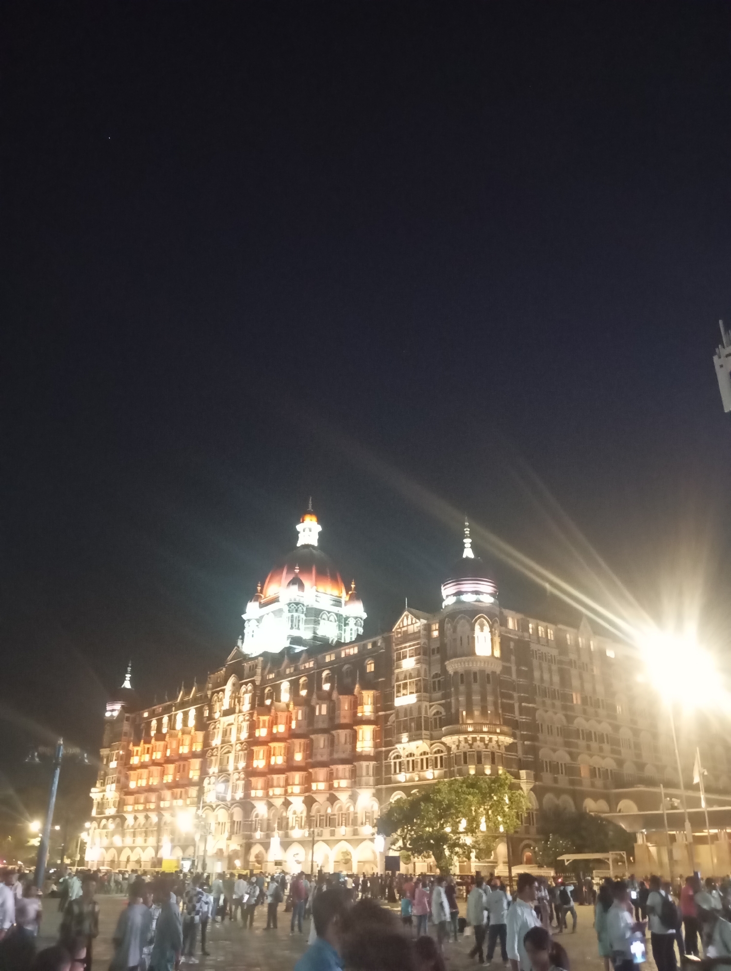 The image depicts the Taj Mahal Palace hotel in Mumbai, India, at night. The building is illuminated with lights, accentuating its architectural details. A crowd of people is gathered in front of the hotel. The sky is dark. There are streetlights illuminating the scene.