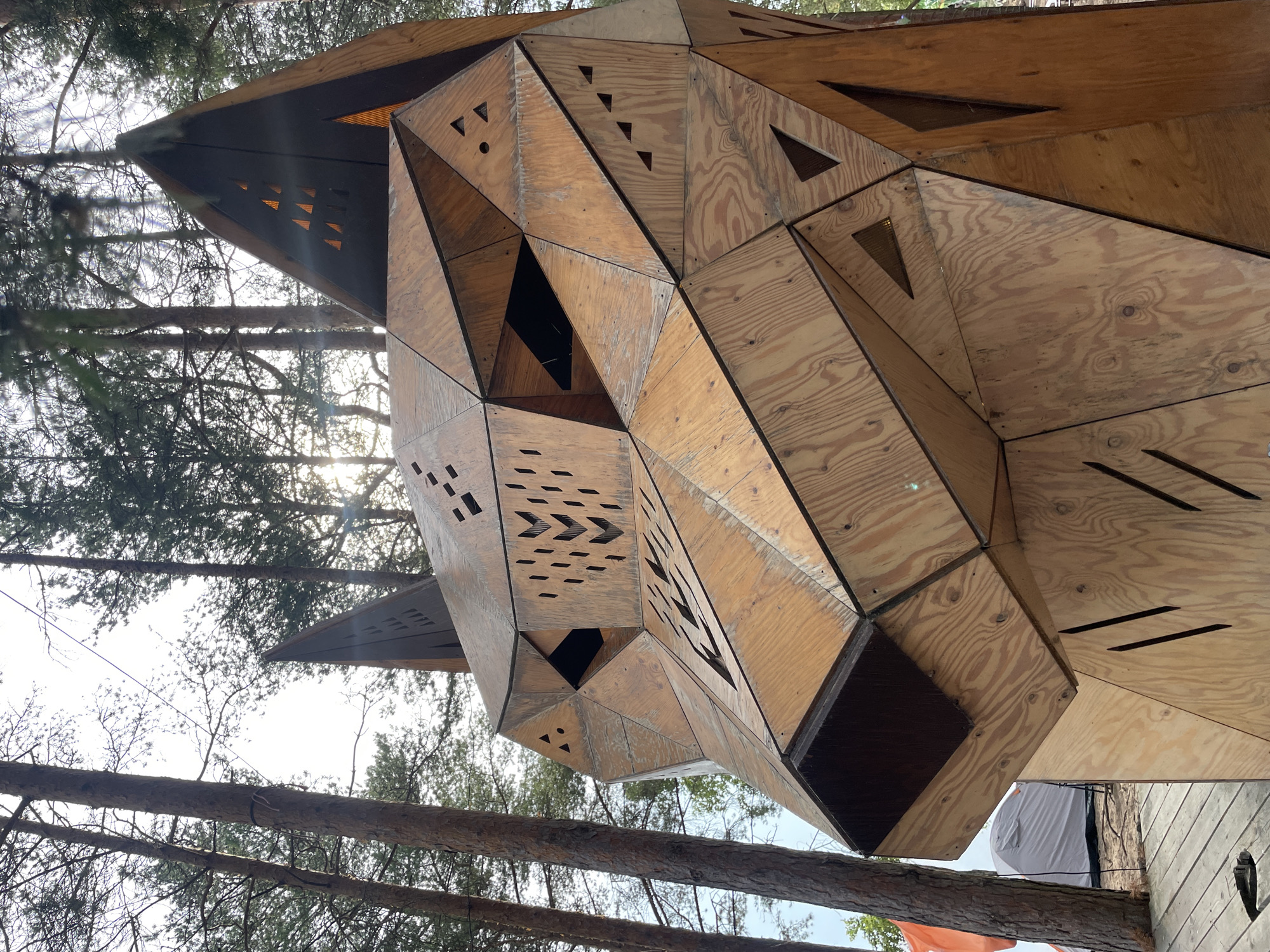 The image shows a large, geometric wooden sculpture of a wolf's head, constructed from plywood panels. The sculpture is located outdoors, in a forest setting with tall pine trees. The sun is visible through the trees, casting dappled light on the sculpture. In the background, there are tents and a wooden deck, suggesting a campsite or festival. The sculpture has intricate details, including cut-out patterns on its face, resembling eyes, nose, and mouth. The wolf's head is depicted in a stylized, angular manner, with sharp lines and planes. The texture of the plywood is visible, with its natural grain and knots. The overall impression is one of a striking art installation within a natural environment. The location is Luzhki, Russia.