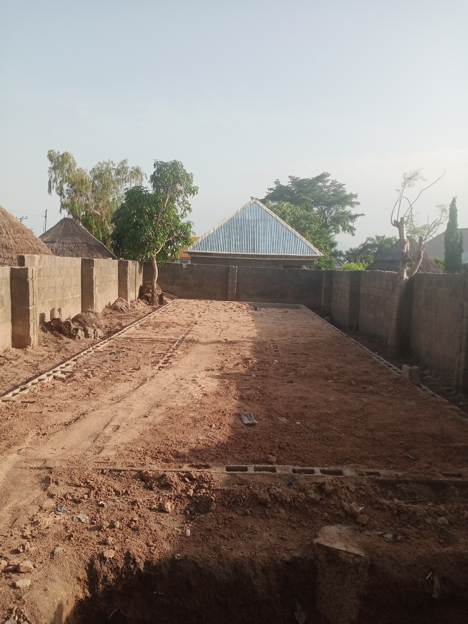 The image depicts an outdoor construction site in Jalingo, Nigeria. The foreground is dominated by a large, flat area of reddish-brown soil, appearing to be a foundation or cleared building plot. Tire tracks are visible in the dirt, suggesting the recent use of vehicles. Along the left and right edges of the image are brick walls, some of which are partially constructed with cinder blocks. In the background, several thatched-roof structures and a building with a corrugated metal roof are visible, along with various trees and vegetation. The sky above is clear and pale blue, indicating daytime. There are no people or animals visible in the image. The scene conveys a sense of ongoing construction and development in a semi-urban or rural setting.