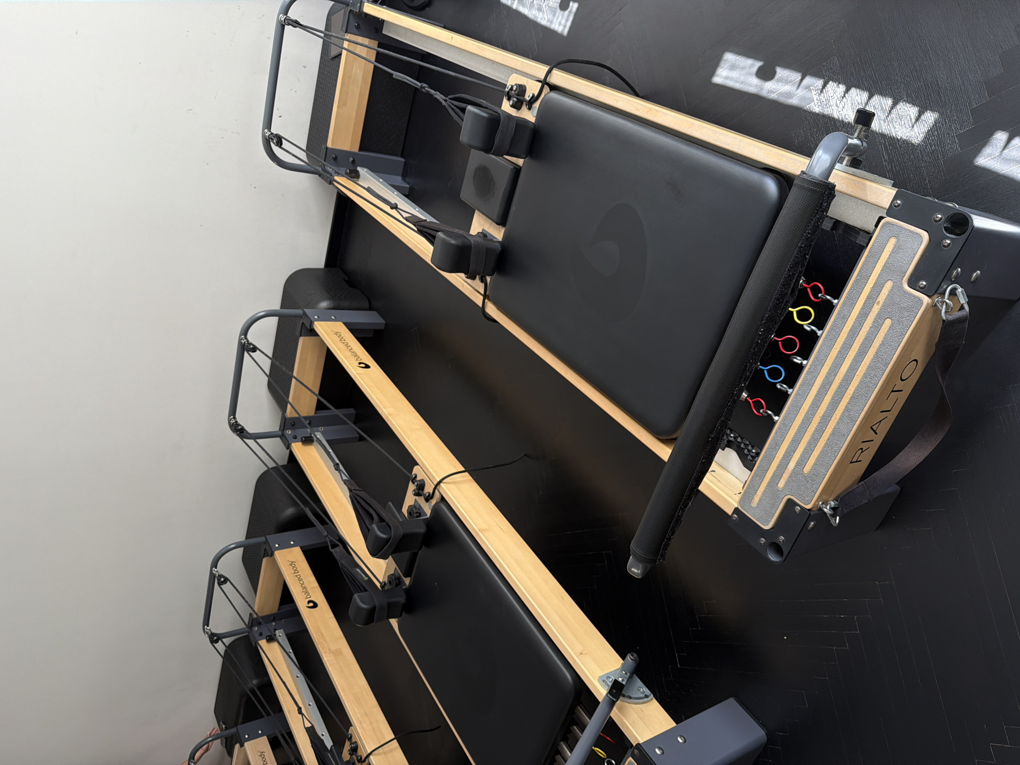 The image shows three Pilates reformers, stacked vertically against a wall. The wall to the left is off-white, while the floor is black with a subtle, herringbone pattern. The reformers are made of light-colored wood and black upholstery. The top reformer is a "Rialto" model, indicated by the name visible on the footbar. Each reformer has adjustable straps, pulleys, and a carriage. The overall scene appears to be a Pilates studio or a storage area for Pilates equipment. The location, Belgrade, Serbia, is not discernible from the visual information provided in the image.