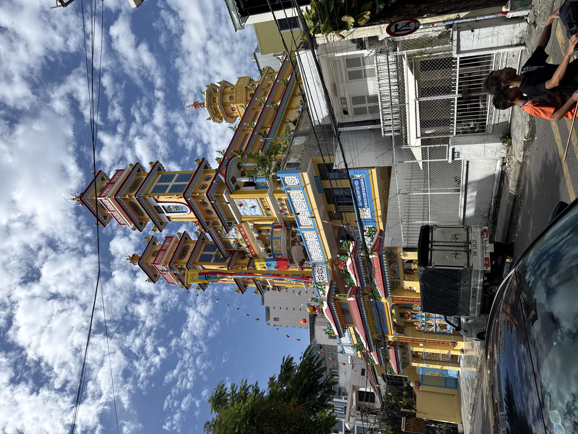 The image captures a colorful Buddhist temple in Nha Trang, Vietnam. The temple is adorned with yellow, red, and blue paint, featuring multiple tiered roofs and decorative statues. The architectural style is distinctly Vietnamese, with intricate carvings and vibrant banners.
In the foreground, a silver truck with a dark tarp cover is parked on the street. The truck has Vietnamese license plates. To the right of the truck, two young boys are riding a motorcycle. The older boy, wearing a black t-shirt, is driving, and the younger boy, in an orange t-shirt, is the passenger. They are on a paved street with yellow pedestrian crossings.
The background shows a dense urban environment with other buildings, trees, and power lines crisscrossing the sky. The sky is blue with fluffy white clouds, suggesting a sunny day. The overall scene depicts a typical street view in a Vietnamese city, with a religious landmark as the central focus.
