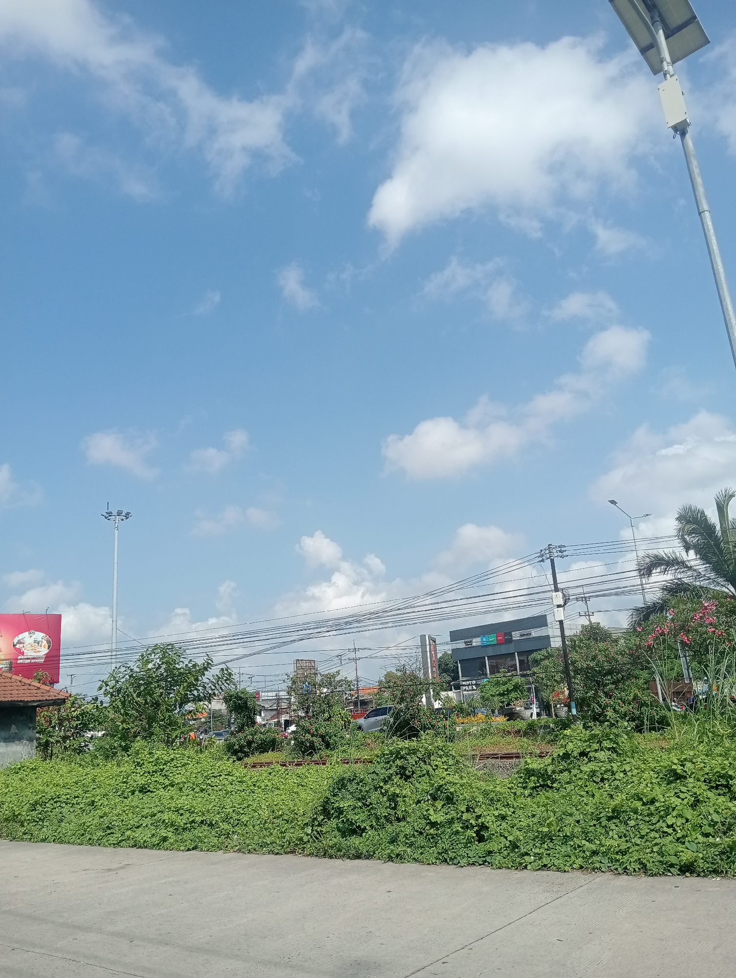 This image captures a bright, sunny day in Sawotratap, Indonesia, with a clear blue sky dotted with white clouds. The foreground shows a paved surface, possibly a road or walkway, bordering a dense area of lush green vegetation. Beyond the vegetation, a line of utility poles with numerous wires stretches across the scene, indicating an urban or semi-urban environment. Several buildings are visible in the mid-ground, including a building with a dark facade and some signage, suggesting commercial activity. A tall light pole with multiple lamps stands to the left, and a solar-powered street lamp is positioned on the right. A billboard with red accents is also present on the left side of the image. The presence of trees and flowering plants, particularly pink flowers on the right, adds to the natural elements within the built environment. The overall scene depicts a typical day in a developed area of Sawotratap.