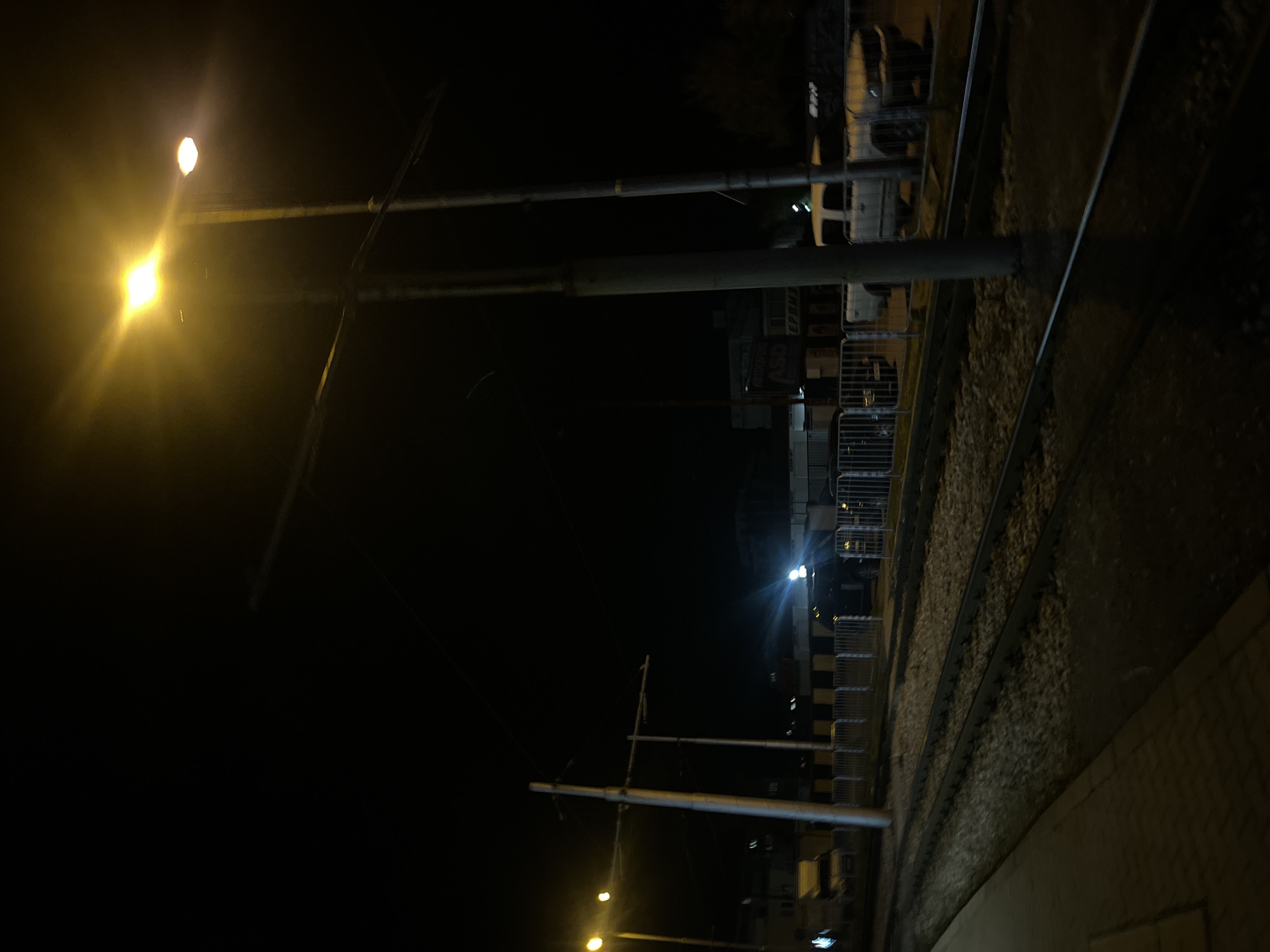 At night, a train track runs along the right side of the frame, with a platform alongside it at the bottom right. The left side of the image is dark, illuminated by streetlights on poles. Some buildings can be seen in the distance, along with a few parked vehicles. The location is likely Sofia, Bulgaria, given the appearance of the infrastructure.