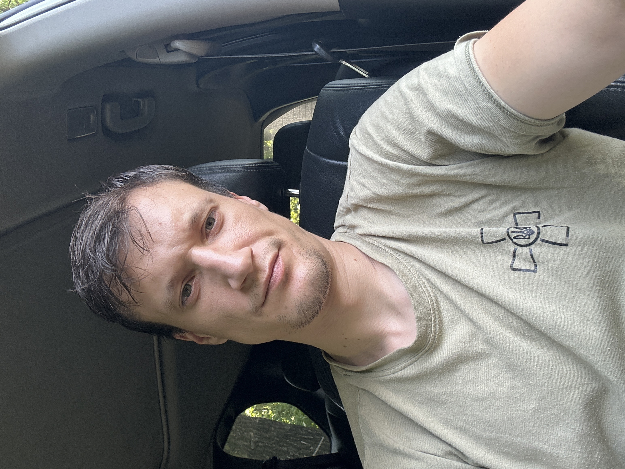 A man is sitting in the driver's seat of a car. He is looking at the camera and has short, dark hair and a light complexion. He is wearing a light green t-shirt with a black emblem on the left side of the chest. The emblem is a cross with a trident in the center. The interior of the car is visible, including the dashboard and seats. The man appears to be in a wooded area, with trees and sunlight visible through the car windows. The location is Makiv, Ukraine.