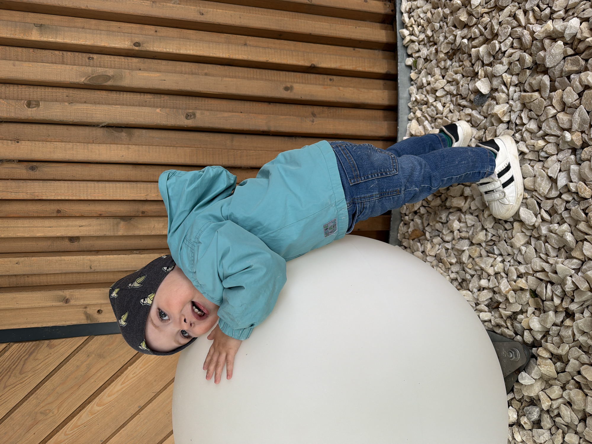 A young child wearing a blue jacket, jeans, a patterned hat, and white sneakers is lying on a white, round structure. The child is looking at the camera with a slight smile. The background consists of light brown wooden planks on one side and a gravel surface on the other. The location appears to be outdoors, but the information provided does not indicate that the location is in Votkinsk, Russia.