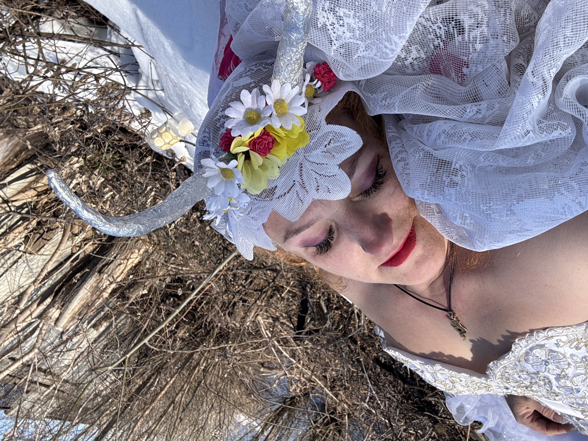 A woman lies supine on a bed of dry vegetation and bare branches, possibly outdoors. She is wearing a white, lacy head covering adorned with silver horns and artificial flowers. She has fair skin, reddish hair, and is wearing red lipstick and pink eyeshadow. She also wears a pendant necklace. She is dressed in a white gown with silver embellishments. The location is presumed to be in Chaikovsky, Russia.