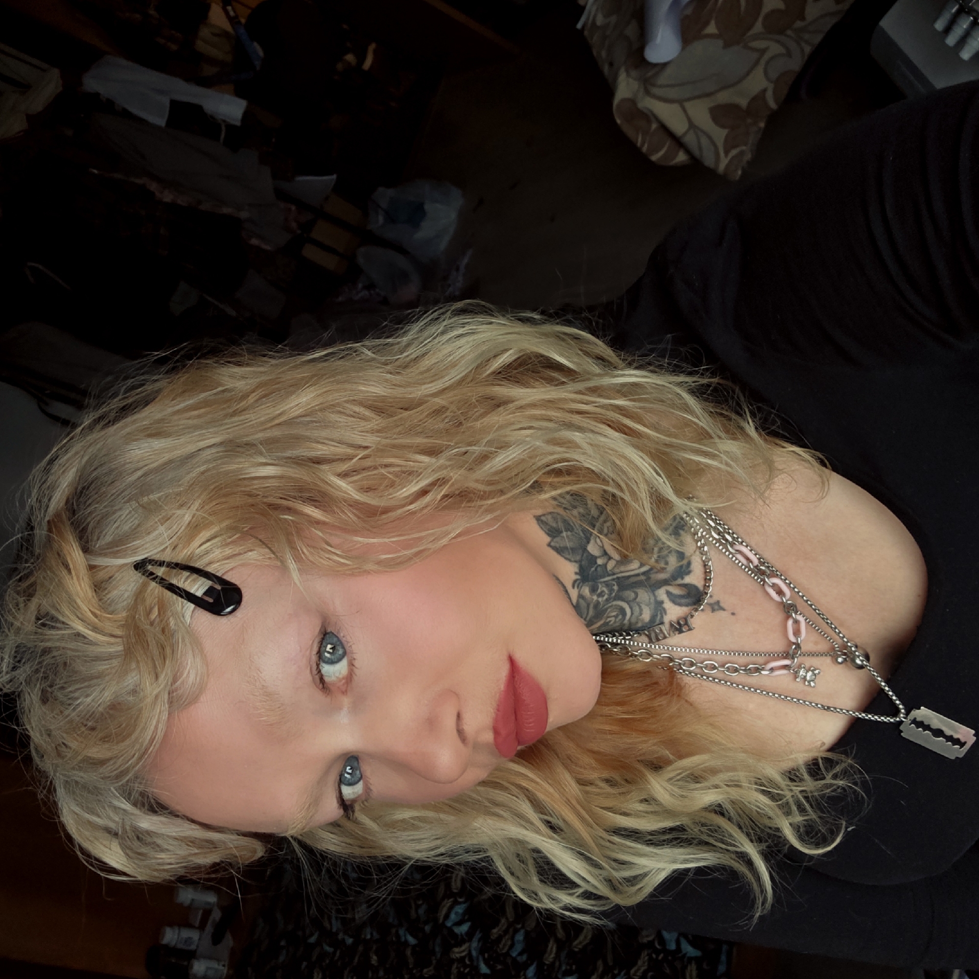 A blonde woman with blue eyes is shown in a close-up selfie. Her hair is wavy and secured with a black clip on one side. She has a neck tattoo and is wearing multiple silver necklaces, one with a razor blade pendant. She wears red lipstick. The background is cluttered with various objects, including a patterned blanket and clothes. The image does not provide information to determine whether the image was taken in Saint Petersburg, Russia.