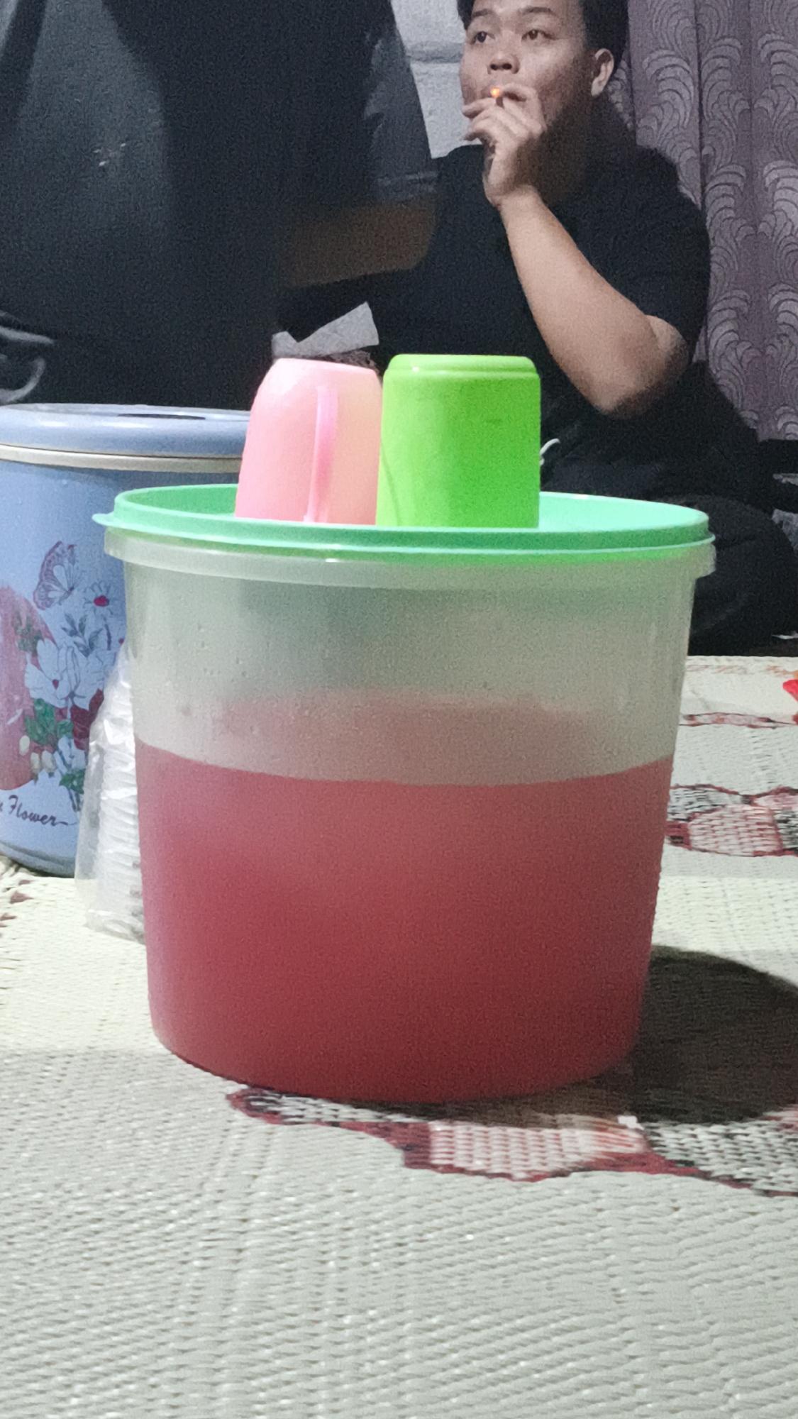 In Kerep Kidul, Indonesia, a close-up shot focuses on a large, translucent plastic pitcher filled with a pink liquid, placed on a woven mat. Two plastic cups, one pink and one green, stand upright inside the pitcher's rim. To the left of the pitcher, a blue bucket with a floral design is partially visible. In the background, a person wearing a black t-shirt is seated, with a cigarette held to their lips, and they are looking to the right. The overall scene is dimly lit, with some objects casting shadows on the mat.