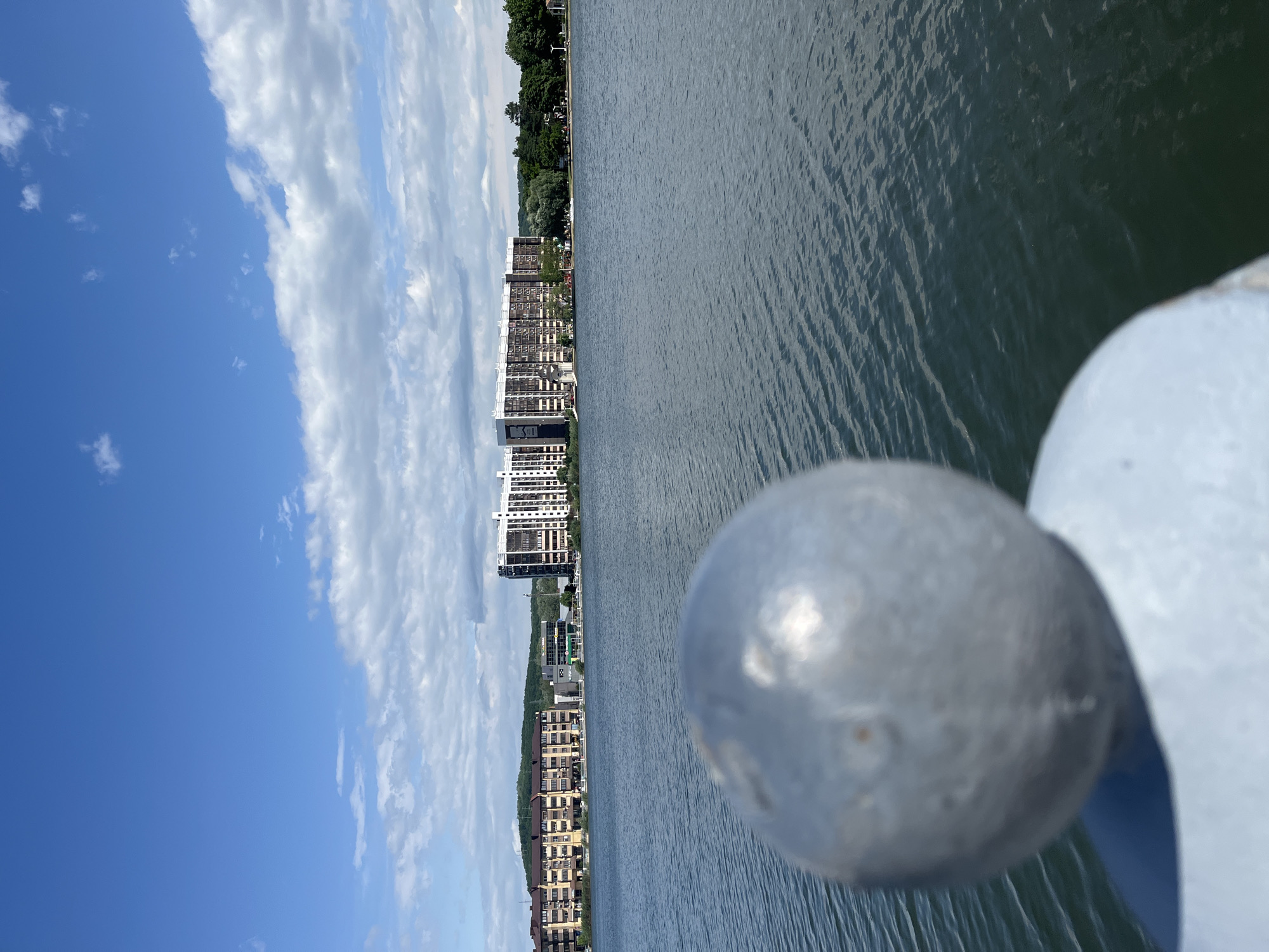The image depicts a view from a pier or bridge in Goryachiy Klyuch, Russia, overlooking a body of water, potentially a river or lake. In the foreground, a metallic sphere, possibly a railing ornament, is visible. The water has a rippled texture, suggesting a slight breeze or current. Along the shoreline opposite the pier, there are several buildings, including what appear to be apartment complexes or hotels. Lush green trees border the buildings. The sky is partly cloudy, with blue sky visible. There are no people visible.