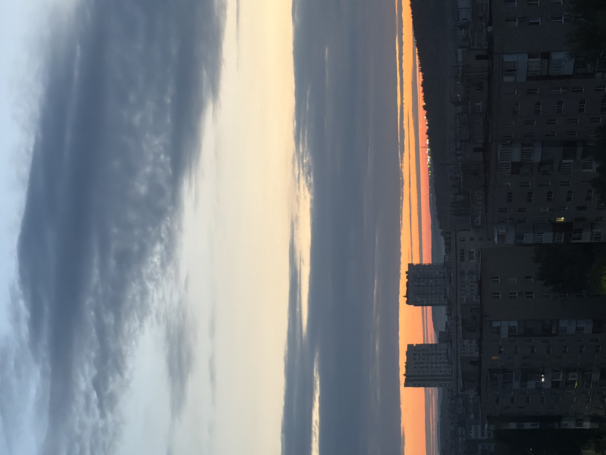 The image shows a cityscape at dusk with a dramatic sky. Two tall, light-colored apartment buildings stand prominently in the mid-ground, silhouetted against a bright orange and yellow horizon. In the foreground and extending to the left and right, are darker, more weathered apartment buildings. The sky is filled with layered clouds in varying shades of gray, blue, and a pale yellow near the horizon. A distant, dark, tree-covered hill or ridge is visible behind the apartment buildings, and a faint line of lights can be seen along its crest. The overall impression is one of an urban environment transitioning from day to night.