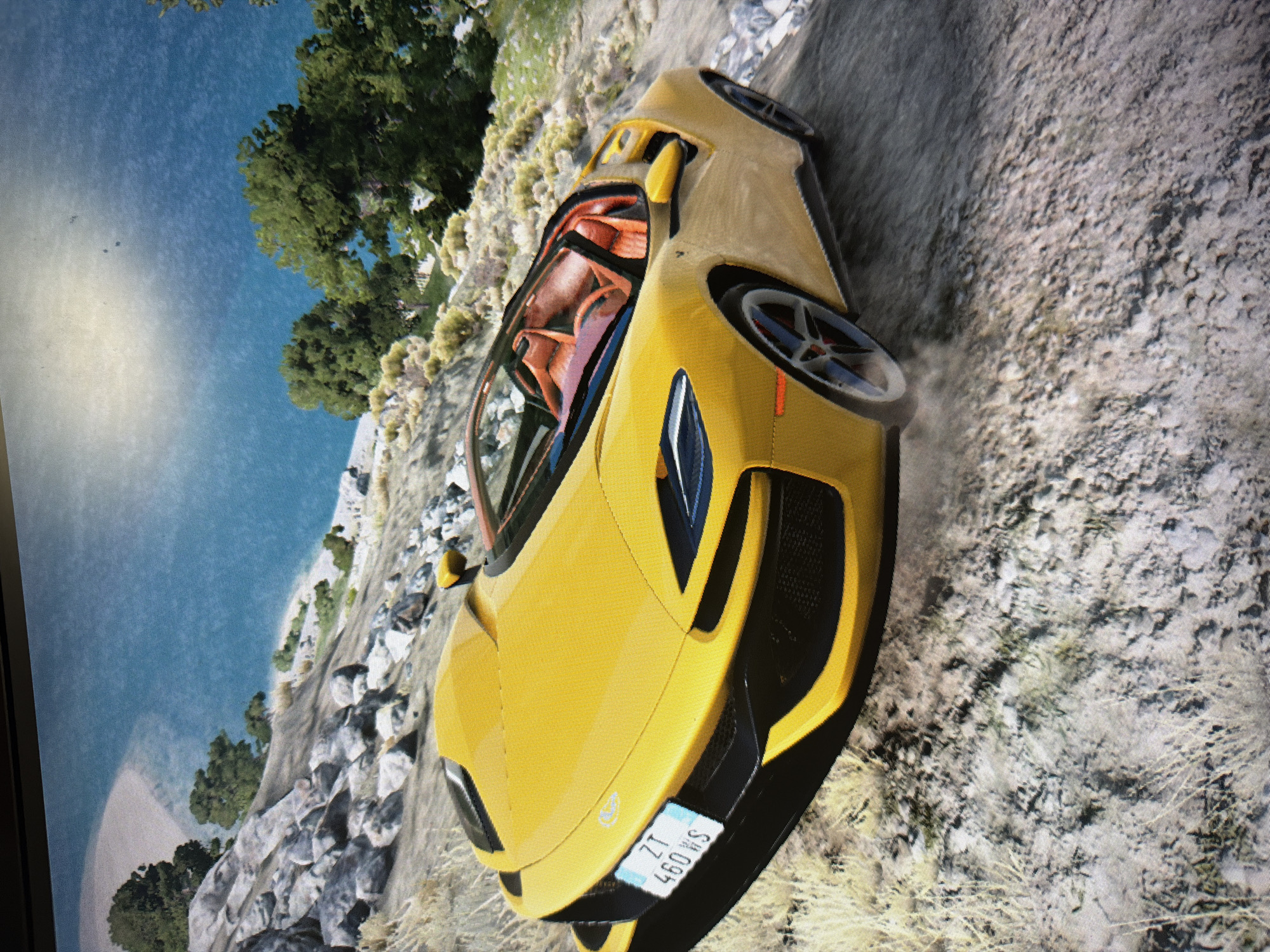 A bright yellow sports car with an orange interior is depicted on a dirt or gravel path in what appears to be a scene from a video game. The image is a photograph of a screen, rotated 90 degrees clockwise, showing visible pixels and some glare on the left side.
The main subject is the sleek, modern sports car, possibly a McLaren model, positioned on an unpaved road that winds along a scenic coastal or lakeside area. The car's front end is prominent, showcasing its distinctive headlights and grille. A subtle cloud of dust near the rear wheel suggests recent movement or acceleration on the loose terrain.
The setting is a vibrant, sunny day with a clear blue sky. The environment includes rocky cliffs, lush green trees, and a vast expanse of blue water to the left. The lighting indicates daytime conditions with no apparent adverse weather. In the upper left corner of the original image (which becomes the top in the rotated view), faint, blurry objects are visible against the sky, possibly distant aircraft or birds.
A white license plate is attached to the front bumper of the car. When viewed with the image rotated to standard orientation, the text on the license plate reads vertically as "SM 097" and "ZT 460 WS". The overall scene evokes a sense of dynamic driving through a picturesque virtual landscape.