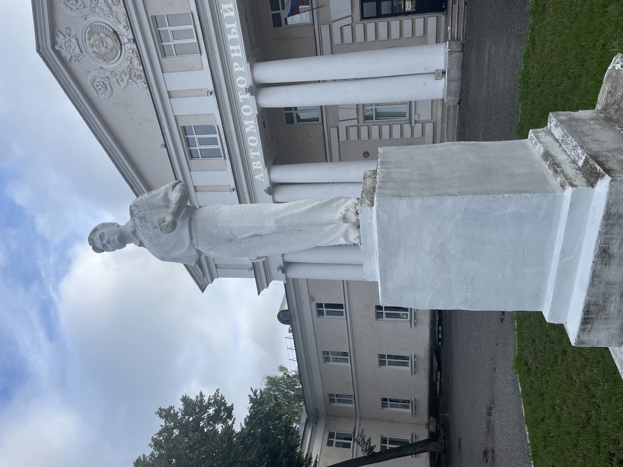 The image shows a white statue of a young man standing on a rectangular pedestal in front of a light-colored building. The building has several windows and large white columns, and there is Russian text above the columns that reads "АВТОМОТОРНЫЙ" (Automotorny). A Russian flag is visible on the right side of the building. The statue is in the foreground, and the building occupies the background. The area around the statue appears to be an outdoor plaza or courtyard, with some grass and a paved surface. The sky is overcast. This scene is located in Zavolzhye, Russia.