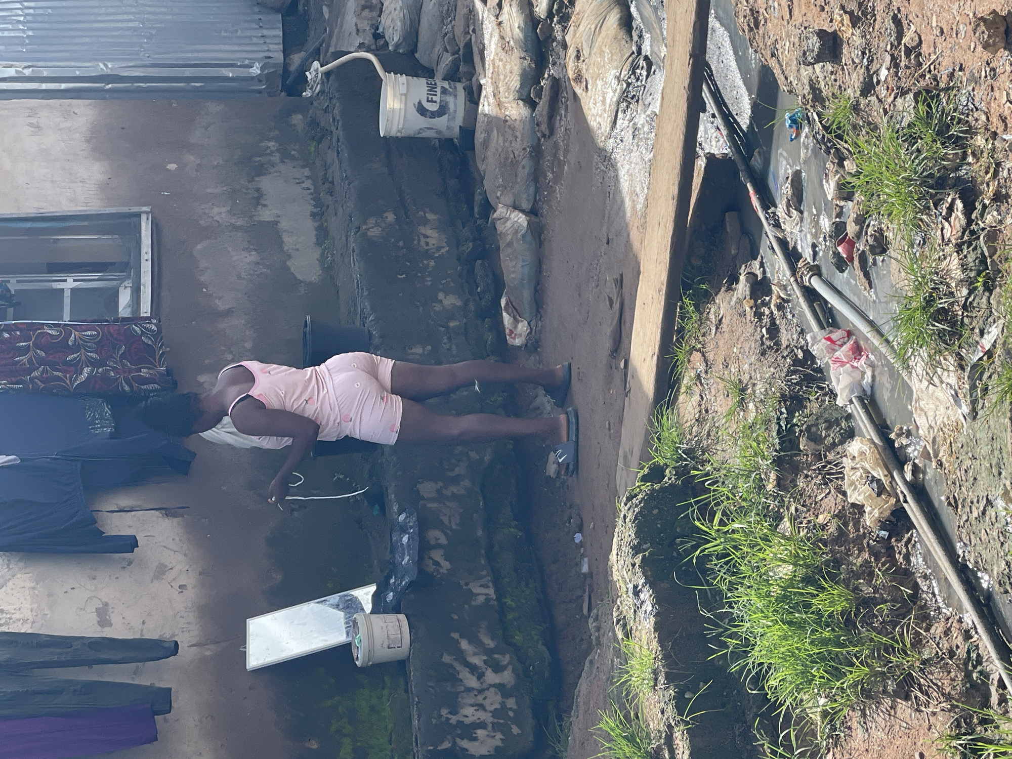 In an outdoor setting that appears to be a residential area in Abuja, Nigeria, a young woman stands near a building with a rough-textured wall. She is wearing light pink pajamas with a peachy-colored print. Her hair is pulled back, and she is holding a white cord in her right hand. A large mirror leans against the wall to her left, and a light-colored bucket is situated in front of it. Clothes hang on a line above the woman. The ground is uneven, with dirt and debris. A section of the foreground shows a small ditch filled with murky water and some debris, including plastic bags and a red cup. A wooden plank lies across the ditch. In the background, a white bucket with the word "FINE" visible on it is near the building. To the far right, corrugated metal sheeting is attached to the building.