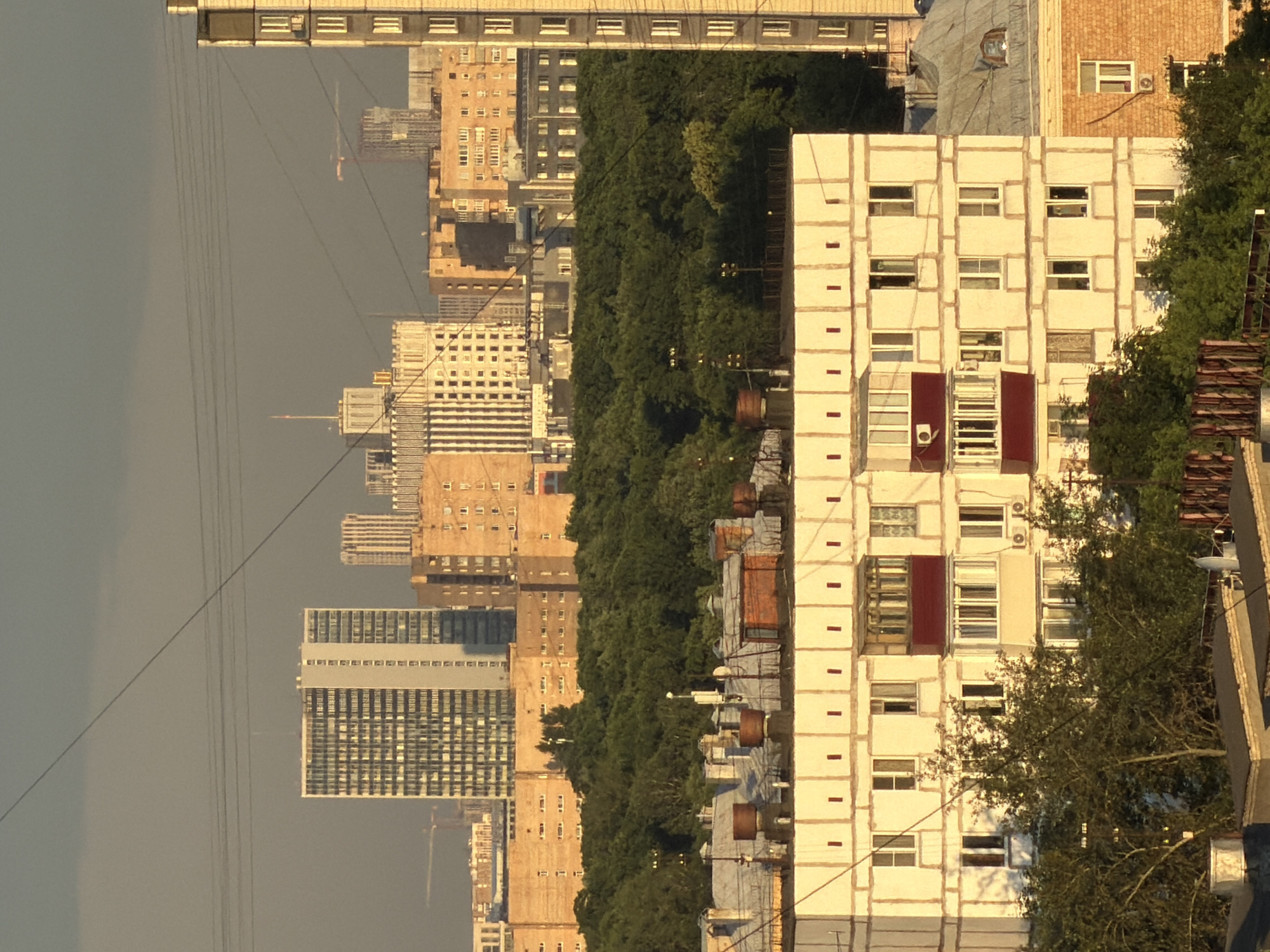 The image captures an urban landscape in Khimki, Russia, under a dusky sky. In the foreground, a light-colored apartment building with a prominent red-trimmed balcony on its third floor is visible. This building features multiple windows, some adorned with air conditioning units. Behind it, a dense expanse of dark green trees stretches across the mid-ground.
Further back, a series of apartment blocks and office buildings rise into the distance. One of these structures is a tall, light-colored building with a grid-like pattern of windows. Another notable building in the background is a larger, light-colored structure with ornate architectural detailing and a flagpole on top, illuminated by the fading sunlight. A crane, potentially indicating ongoing construction, is also visible in the middle distance. The scene is crisscrossed by overhead wires. The overall impression is of a densely populated urban area with a mix of residential and commercial architecture, partially enveloped by greenery.