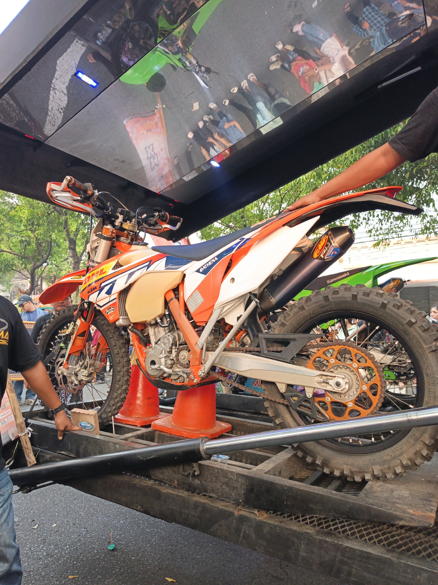 An orange and white dirt bike is positioned on a black trailer. The bike is angled to the right, with its front wheel slightly elevated. A person's arm, wearing a black short-sleeved shirt, is seen resting on the rear fender of the motorcycle. In the background, several people are visible, some standing and others reflected in a dark, reflective surface above the trailer. There are trees and buildings in the distance. The ground beneath the trailer is asphalt. The scene appears to be outdoors, possibly during a public event, given the presence of people and the trailer setup. The location is identified as Gayamprit, Indonesia.