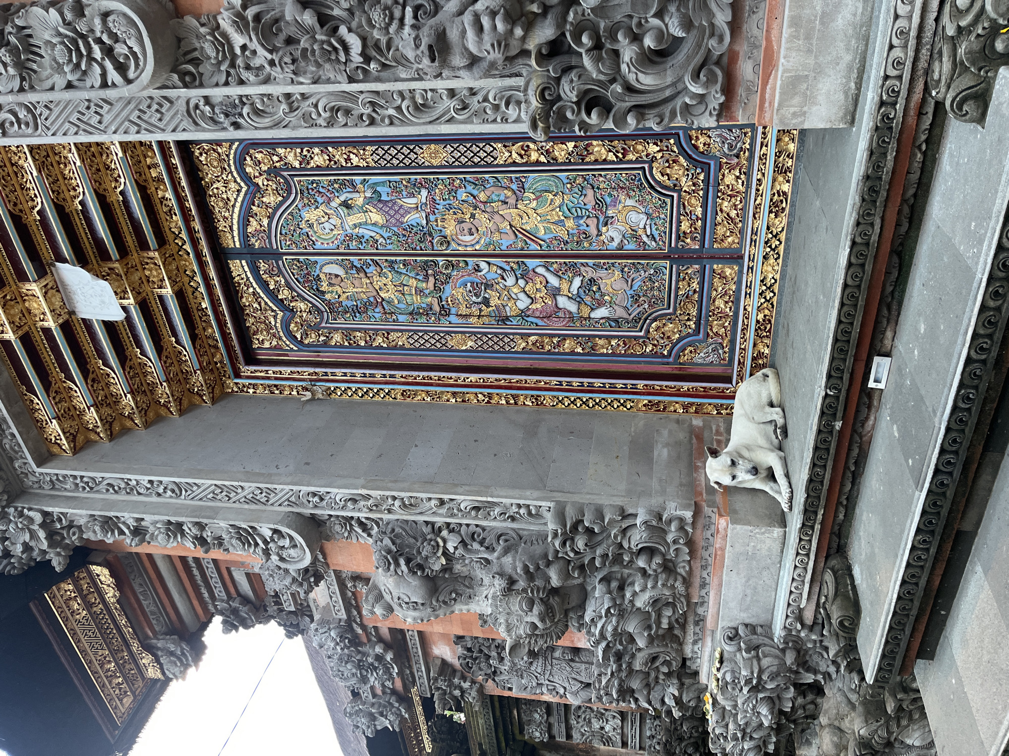 A low-angle, vertically oriented photograph depicts the interior of a temple structure in the Ubud District, Indonesia. The temple's architecture showcases elaborate stone carvings and painted ceilings. A cream-colored dog is lying on a ledge. The ceilings are adorned with intricate painted panels featuring figures and ornate gold detailing. Carved stone elements include floral and mythical creature motifs. The overall environment suggests a blend of artistic detail and traditional construction, typical of Balinese temple architecture.