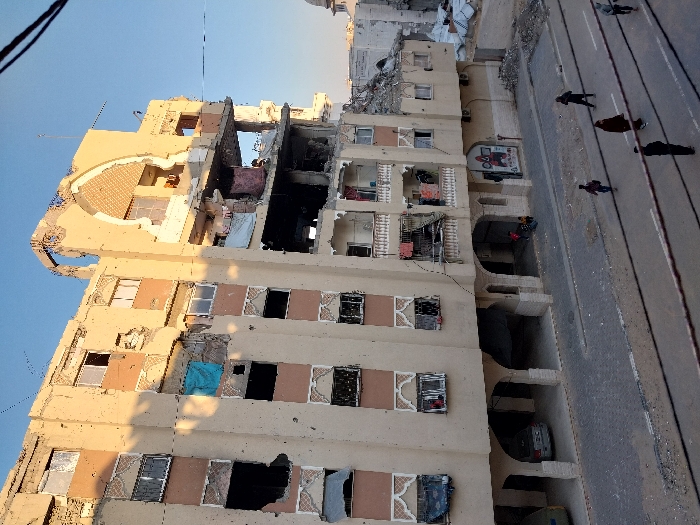 The image displays a multi-story residential building in Khan Yunis, Palestinian Territory, exhibiting significant structural damage under a clear sky. The building, with a beige and light brown facade featuring decorative elements around windows, has extensive destruction on its upper floors, particularly on the right side. Large sections of walls are missing, exposing interiors with debris and twisted rebar. Numerous small impact marks are visible across the entire facade, and many windows are shattered or absent. A distinctive arch-shaped architectural feature on the upper right is partially demolished. Laundry or fabric items hang from balconies and exposed areas.
The ground floor features an arched colonnade where two vehicles are parked, and two children are visible near an entrance. A paved street runs in the foreground, showing scattered debris. Six individuals, dressed in dark clothing, are walking on the street. Overhead wires traverse the upper portion of the frame. In the background, to the right of the main building, further rubble and a portion of a white-domed structure, possibly a mosque, are visible.
