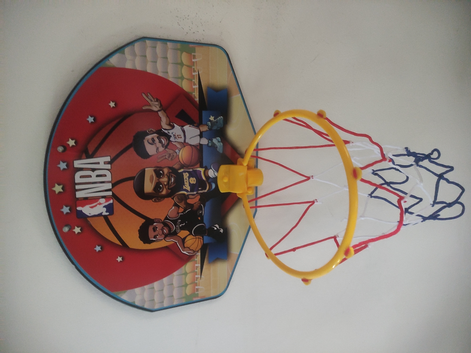 A toy basketball hoop with an NBA-themed backboard is mounted on a light-colored wall. The backboard features cartoon caricatures of three basketball players, a large basketball graphic, and the NBA logo. The hoop itself is yellow, with a red, white, and blue net. The image is shot from a slightly upward angle. The location is the city of Kalisampurno, Indonesia.
