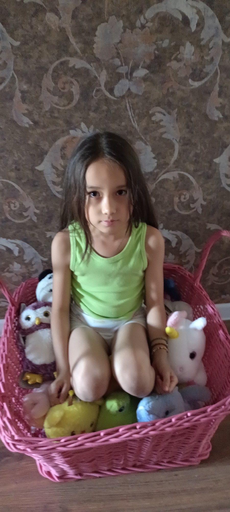 A young girl with long, dark hair is sitting in a large, pink woven basket filled with stuffed toys. She is wearing a light green sleeveless shirt and white shorts. The basket is placed in front of a wall with a brown, floral patterned wallpaper. Among the stuffed toys are an owl, a unicorn, and several small chick-like figures in yellow and green. The girl is looking directly at the camera with a neutral expression. The scene appears to be indoors, likely in a residential setting.