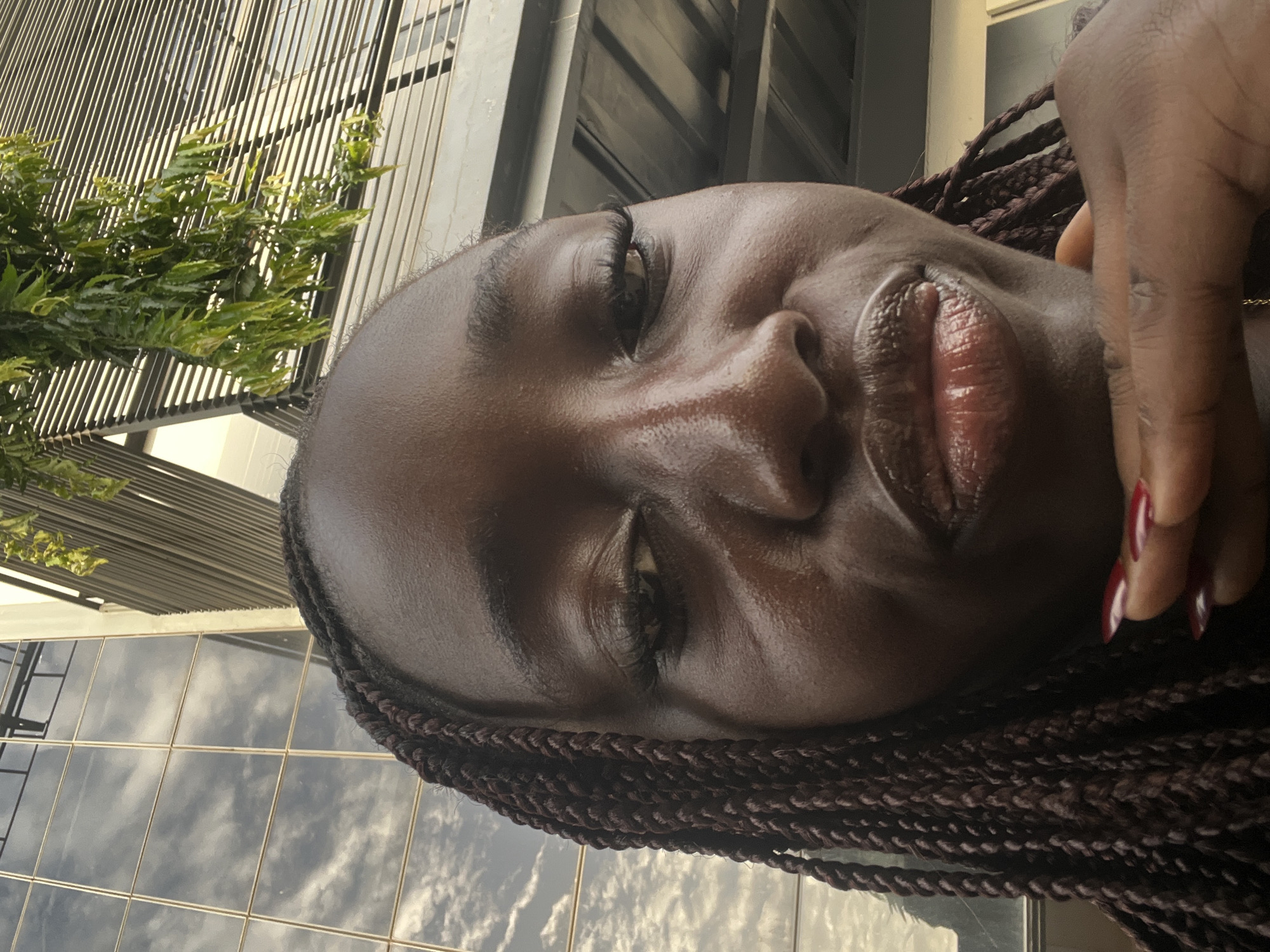 A close-up, slightly tilted shot of a dark-skinned woman's face, with a partial view of her braided hair. Her skin appears moisturized or slightly oily. She is smiling gently, with full, glossy lips. Her eyes are partially closed. Part of a hand with red nail polish is visible near her face. The background includes a building with a glass facade reflecting the sky and clouds. There is also some green foliage visible on the left side of the frame. The location is suggested to be Karum, Nigeria.