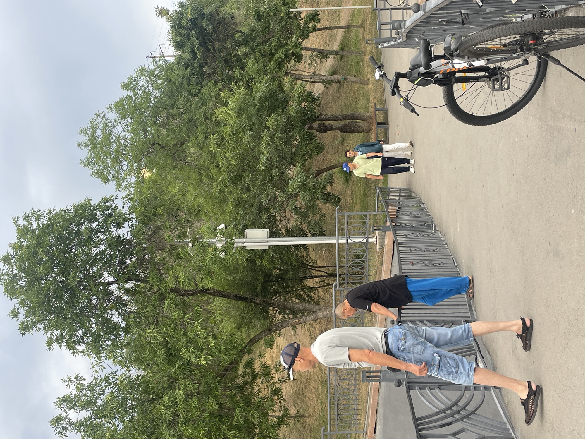 The image depicts an outdoor scene on a paved walkway, possibly a park or promenade, with a metal railing running along its edge. Several people are present. In the foreground, a man wearing a grey t-shirt and denim shorts is walking away from the camera. To his left, another man in a black t-shirt and blue trousers leans on the railing. Further down the walkway, two individuals, one in a light green polo shirt and dark trousers, and the other in a blue jacket and white trousers, are walking towards the background. A bicycle is parked near the railing on the right side of the image. Lush green trees and a sloping grassy embankment form the background, with a cloudy sky overhead. Based on the presence of these individuals and the urban park-like setting, this scene is consistent with a public space in Almaty, Kazakhstan, where people commonly engage in outdoor activities.