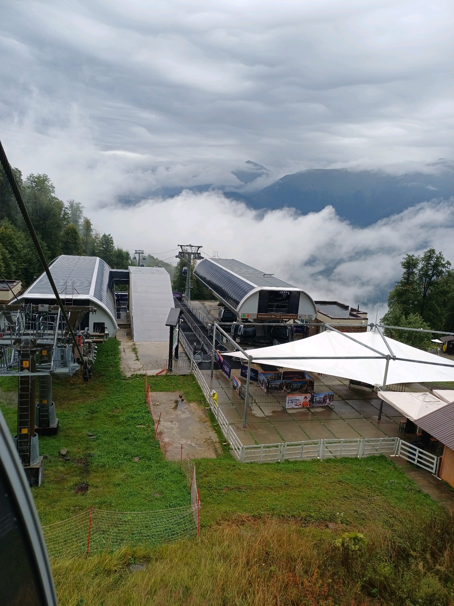 The image depicts the base station of a cable car in Estosadok, Russia, set against a backdrop of misty mountains. The cable car station features two main structures: a modern, curved building with a translucent roof and a more utilitarian white structure. A large, dark-colored cable car cabin is visible at the station. In front of one of the buildings, there is a white tented area covering a waiting or seating area with banners displayed. Several other cable car towers and lines extend into the distance, disappearing into the fog. The surrounding environment is characterized by lush green grass, trees on the slopes, and a cloudy, overcast sky. A paved walkway and a fence are also present. A person wearing a yellow jacket stands near the fence, and other indistinct figures are visible in the background. The scene suggests a mountain resort environment, likely during a cloudy or rainy day given the mist and wet ground.