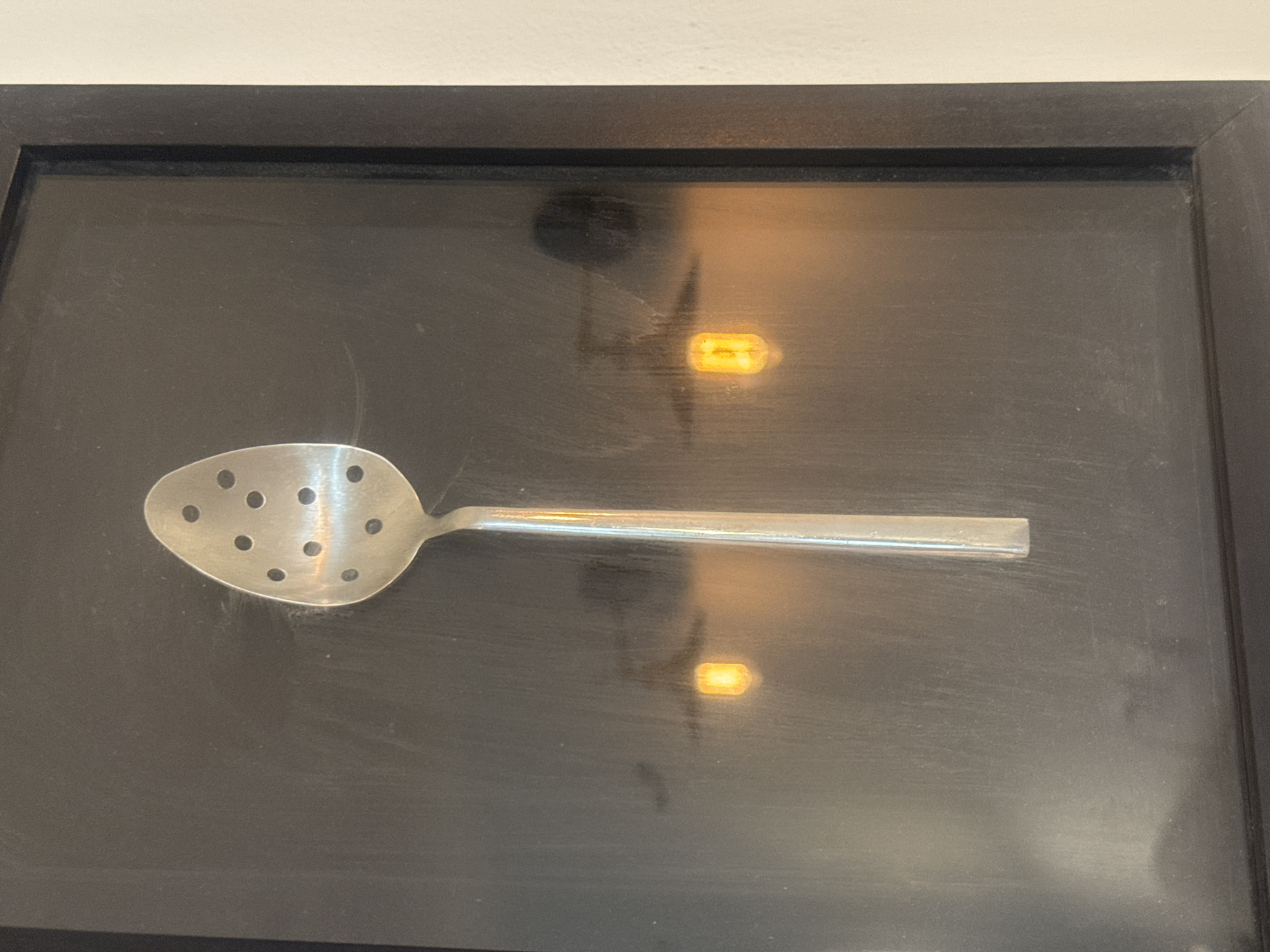 A silver slotted spoon lies in a black-framed display. The display sits against an off-white wall. Reflections are visible on the glass covering the display. No information is available about the city of Tel-Aviv, Israel.