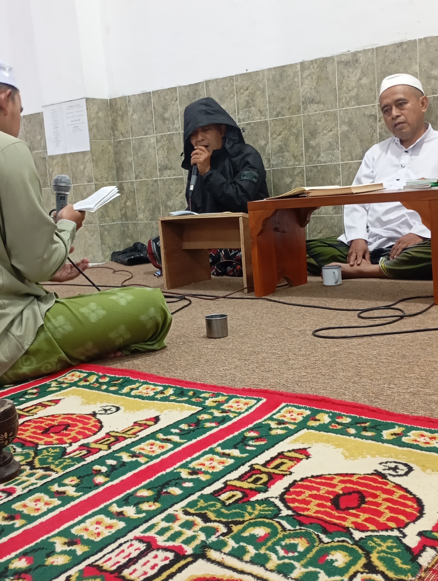 The image depicts an indoor scene where three individuals appear to be engaged in a religious gathering. The room has tiled walls and a carpeted floor. In the foreground, a colorful rug with geometric and symbolic patterns is visible.
One person, wearing a light green traditional outfit and a white cap, is seated on the left, holding a book and a microphone. They are facing away from the camera and appear to be reading or speaking.
In the middle, a person wearing a black hooded jacket is also seated and speaking into a microphone, positioned close to their face. The hood is up, obscuring most of their features.
On the right, a person in a white shirt and a white cap is seated behind a low wooden table. This individual is looking towards the person on the left. The table has a book open on it.
Other objects in the scene include electrical cords on the floor, a small metal cup, and a dark wooden decorative item in the lower left corner. The lighting appears to be from overhead sources, creating a somewhat dim ambiance.
The context suggests a religious recitation or sermon taking place in a community space, potentially a prayer room or a modest hall within the city of Ngadimulyo, Indonesia.
