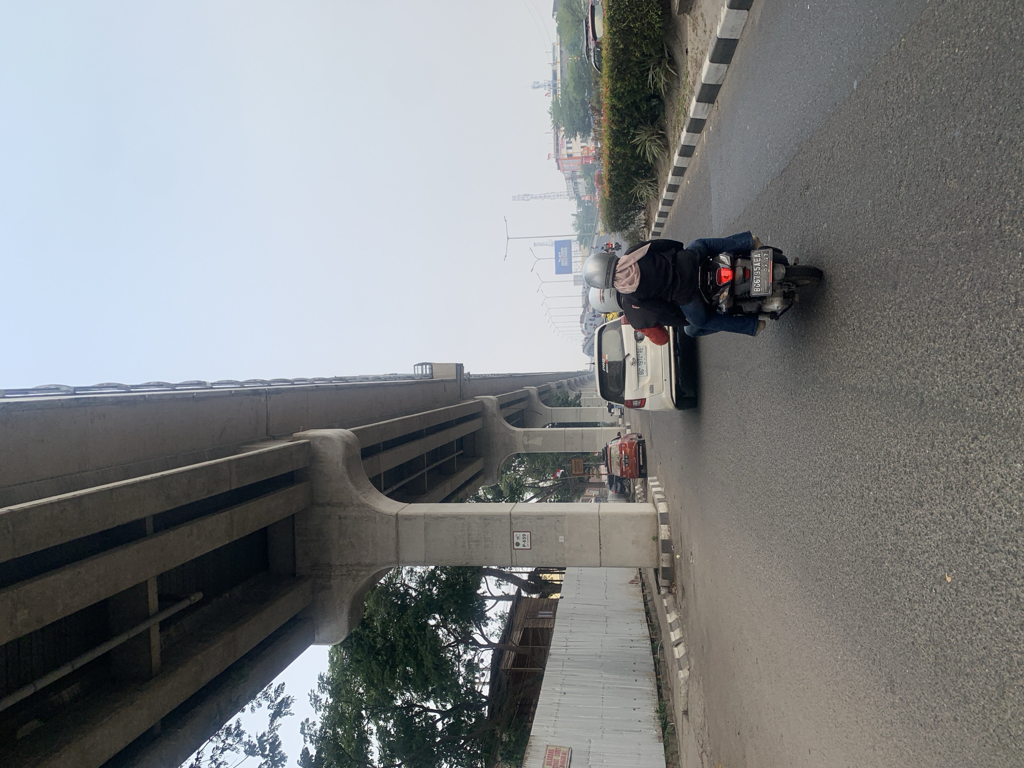 The image captures a street scene in Palembang, Indonesia, with a prominent elevated concrete structure, likely part of an overpass or bridge, dominating the upper portion. The sky is overcast, suggesting hazy or diffused daylight.
In the foreground, a motorcycle carrying two individuals is moving along the asphalt road. The person in the back, wearing a light pink headscarf, is facing away from the camera. The driver is also facing forward. They are traveling in the same direction as other vehicles visible on the road.
To the left of the road, there is a corrugated metal fence, behind which trees and some rustic structures are visible. The concrete pillars supporting the elevated structure are also a significant feature, with signage attached to one of them.
Further down the road, a line of traffic is present, including a white car in the immediate vicinity of the motorcycle, and further back, an orange car. On the right side of the road, a neatly trimmed hedge runs along the pavement, with more distant buildings and traffic lights visible in the background. The overall impression is one of urban infrastructure and daily commuting.