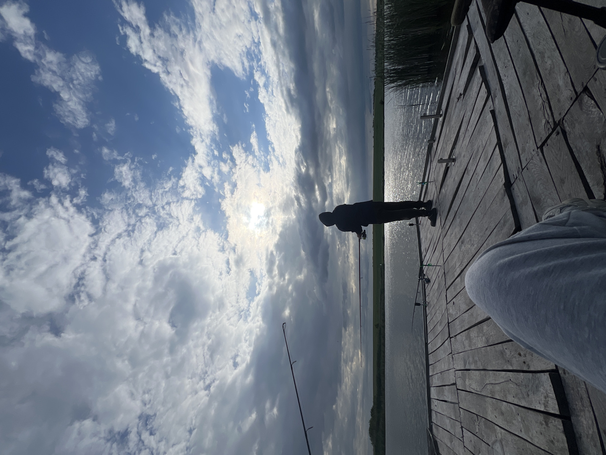 The image depicts a person fishing on a wooden dock. The person is silhouetted against the bright sky, which is filled with clouds. Several fishing rods are visible on the dock. The location appears to be a body of water near some reeds or vegetation. Although the image is presented rotated, the setting seems to be a body of water with a dock at Kopychyntsi, Ukraine.