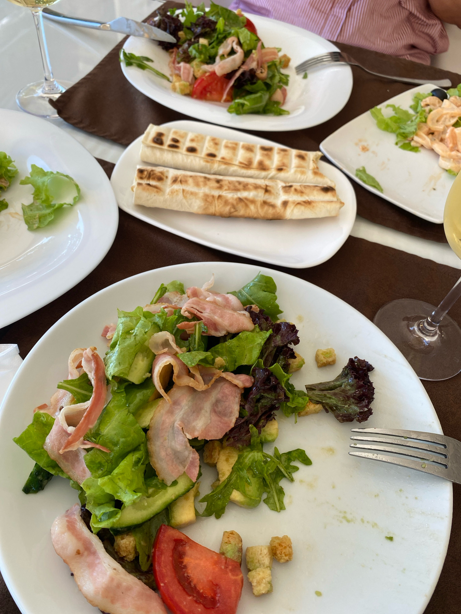 A close-up, high-angle shot shows a table setting for a meal in Chornomorsk, Ukraine. The table is covered with a brown tablecloth. On the table are several white plates containing food, cutlery, and wine glasses.
In the foreground, a white plate holds a salad composed of lettuce, bacon strips, cucumber slices, tomato wedges, and croutons. A fork rests on the edge of this plate.
To the right of the foreground plate, another white plate contains what appears to be a salad with lettuce, mushrooms, and olives. A glass of white wine is visible to the far right.
Behind the foreground plate, a white plate holds two rolled-up flatbreads.
Further back, another white plate displays a salad with lettuce, ham, and tomato. A fork and knife are on this plate, and a wine glass is to the left.
The image depicts a dining scene, likely at a restaurant or a home, with a partially consumed meal. The lighting is bright, suggesting it is daytime.