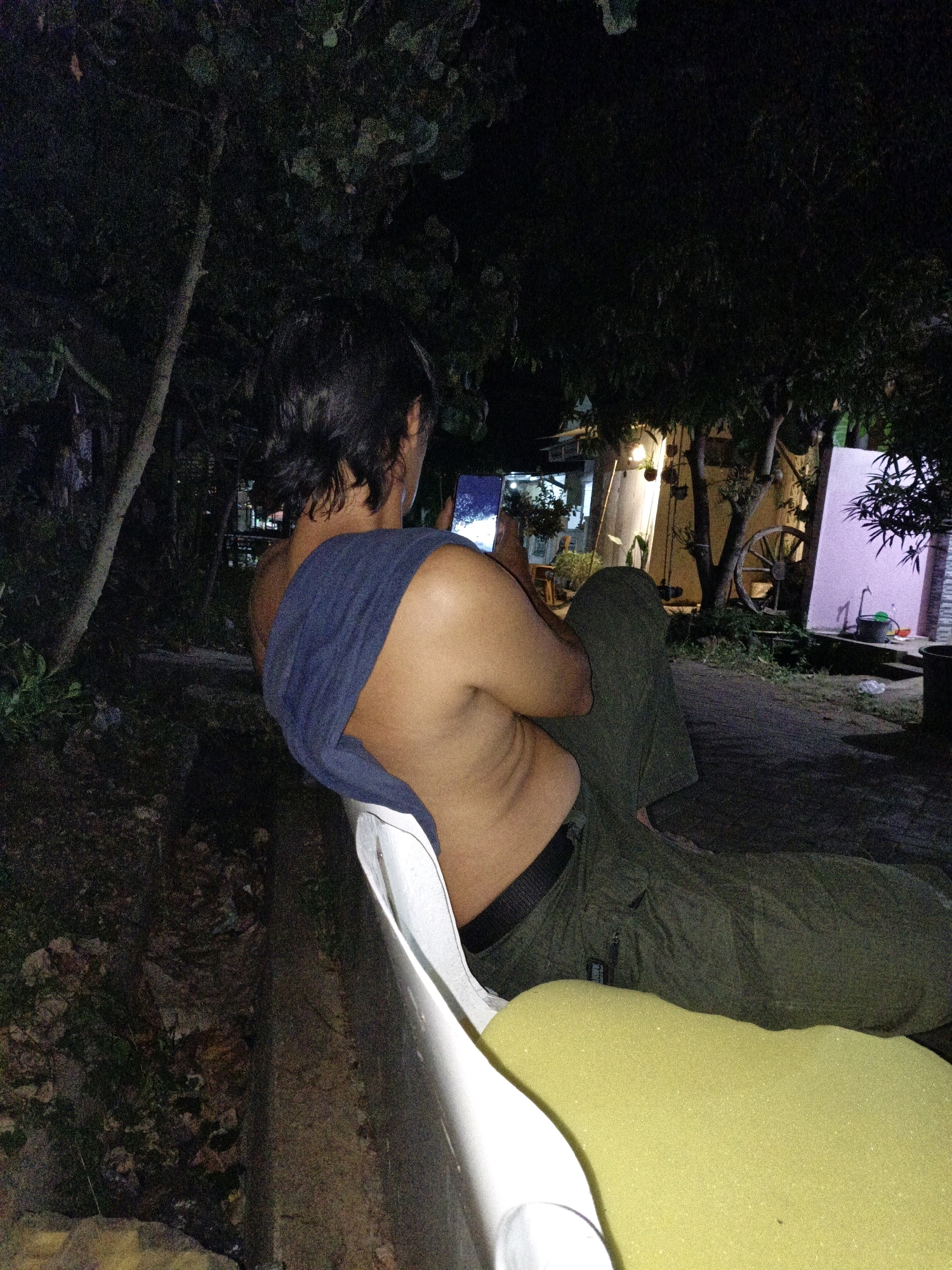A person is sitting on a white chair at night. The person is wearing green pants and has a blue cloth draped over their shoulders. They are looking at a phone in their hands. The person is outdoors, with trees and foliage visible in the background. A building with lights on is also visible in the background. The ground is paved.