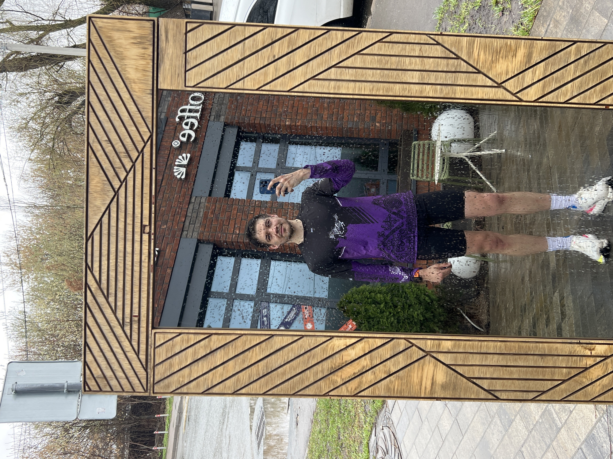 A mirror in Kazan, Russia, reflecting a man in a purple and black shirt and black shorts taking a selfie with a blue phone. The reflection includes a coffee shop facade, outdoor seating, and raindrops on the mirror's surface. The mirror has a wooden frame with a geometric design. The surroundings show street signs, trees, pavement, and grass.