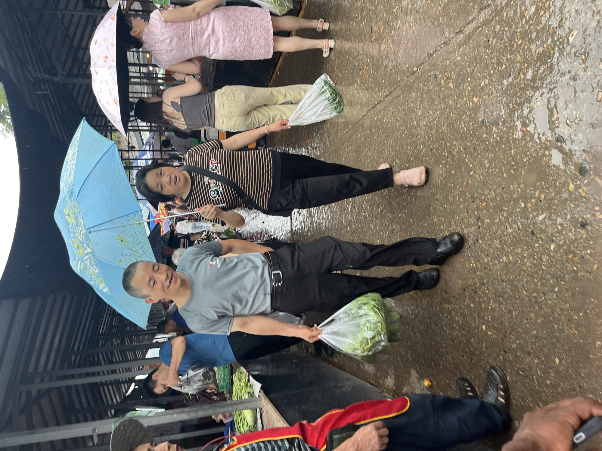 The image captures a lively outdoor market scene in Jiangjin, China, during a period of recent or ongoing rainfall, evidenced by the wet, speckled concrete ground containing numerous puddles and reflective surfaces. An overhead structure, comprising a dark, possibly metal roof supported by metal beams, provides partial cover for the market stalls.
In the central mid-ground, a middle-aged East Asian man with a short, buzzed haircut, dressed in a plain grey t-shirt featuring a small red logo, black pants, and dark dress shoes, walks forward with a broad smile. He holds a clear plastic bag filled with green leafy vegetables in his right hand. Slightly to his right and behind him, a middle-aged East Asian woman walks, holding an open bright blue umbrella with yellow floral patterns over her head for protection from the rain. She is clad in a black and beige horizontally striped t-shirt, black pants, and light pink slip-on sandals, with a black bag slung over her left shoulder and a clear plastic bag of green vegetables carried in her left hand.
To the far left in the foreground, partial views include an individual wearing a straw hat and another wearing a red and yellow striped apron over dark clothing, with dark, perforated shoes, whose hand holds a dark object resembling a smartphone.
In the background on the left, a younger East Asian man in a blue t-shirt is observed interacting with a market stall piled high with green vegetables. Further to the right in the background, an East Asian woman, facing away from the viewer, wears a grey sleeveless top and light-colored pants, carrying a plastic bag. Adjacent to her, another East Asian woman, also facing away, is dressed in a pink patterned short-sleeved dress (resembling a cheongsam) and white sandals, holding a white umbrella with light orange patterns over her head and a plastic bag in her left hand. Numerous other blurry figures are visible throughout the background, indicating a busy public market atmosphere under an overcast sky.