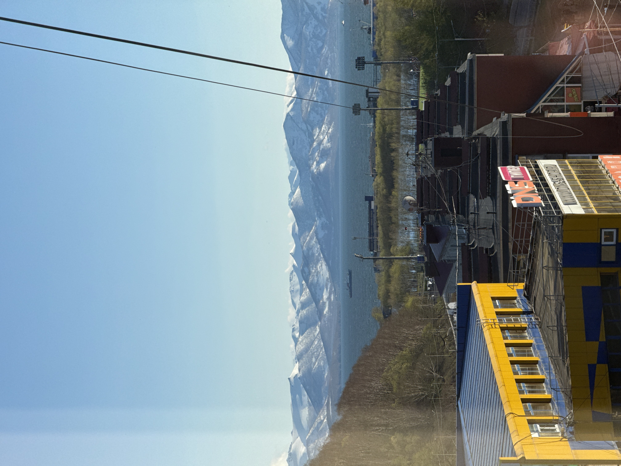 The image captures a view in Petropavlovsk-Kamchatsky, Russia. The foreground shows modern buildings, one with a yellow and blue facade. A brown-brick building and overhead wires also appear. In the background, a body of water stretches towards a mountain range with snow-capped peaks. The sky is clear and blue.