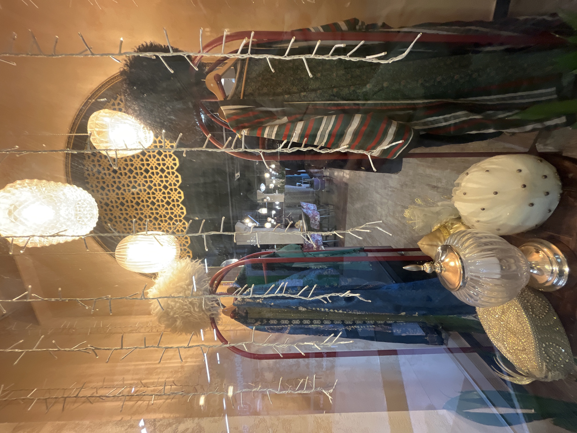 This image depicts a shop window display, photographed from the outside and rotated 90 degrees counter-clockwise. The scene is brightly lit, suggesting an evening or nighttime setting.
A dense network of white fairy lights is strung horizontally across the foreground, partially obscuring the view into the display due to reflections on the glass.
Within the shop window, several items are showcased. On the left side, three brightly illuminated, ornate spherical light fixtures cast a warm glow. Behind them, a golden decorative screen with an intricate, lattice-like pattern adds a distinctive aesthetic.
Towards the center, a red metal display rack holds two garments. The upper garment is a dark, possibly patterned, robe or dress, adorned with a striped shawl featuring red, white, and dark green bands. Below it, a blue-green garment, possibly embellished with intricate patterns, is also displayed.
On the right side of the display, various decorative objects are arranged. These include a fluffy white pom-pom or ornament, a large, round, white textured object studded with dark accents, a clear glass jar with a silver lid and base, and a shimmering gold, leaf-shaped decorative item.
Faint reflections on the window glass reveal glimpses of an interior space beyond the shop display, with visible ceiling lights and indistinct shapes that suggest furniture, potentially indicating a mall or a larger interior setting. There is no legible text visible in the image.