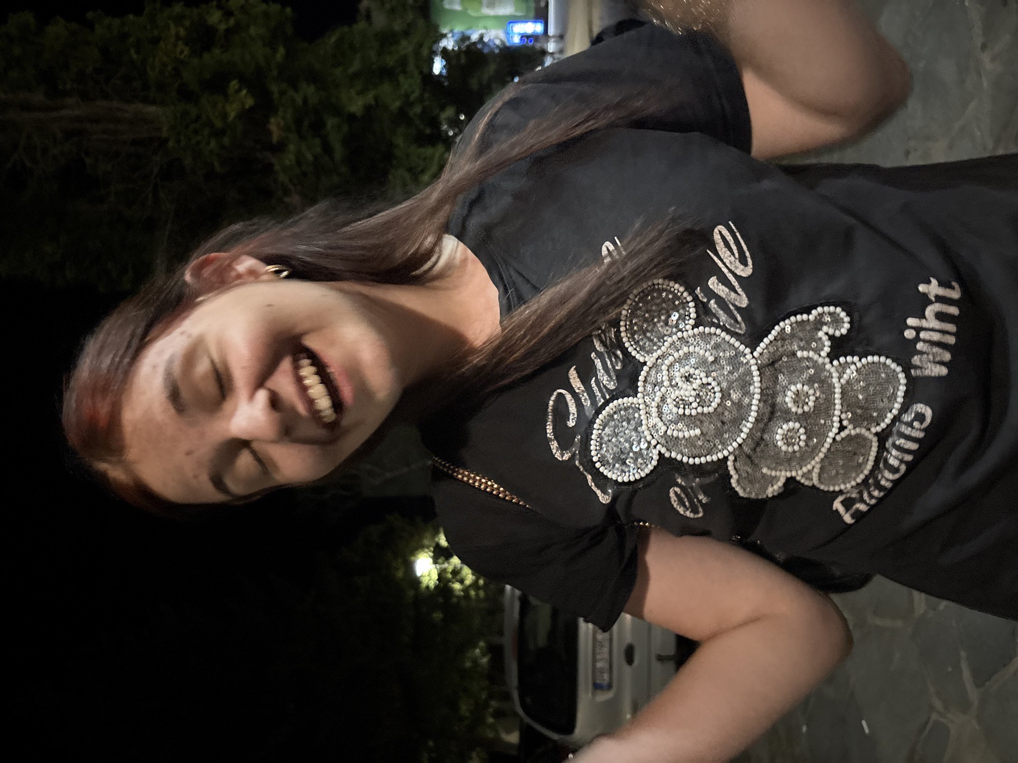 A young woman with auburn hair and freckled skin is pictured at night in Sopot, Bulgaria. She is wearing a black t-shirt adorned with a sequined image of a teddy bear and text. Her eyes are closed, and she is smiling broadly, with her mouth open. She has a gold hoop earring in her visible ear. The background shows trees and the blurred lights of a city at night, with a car parked on the left side of the frame.
