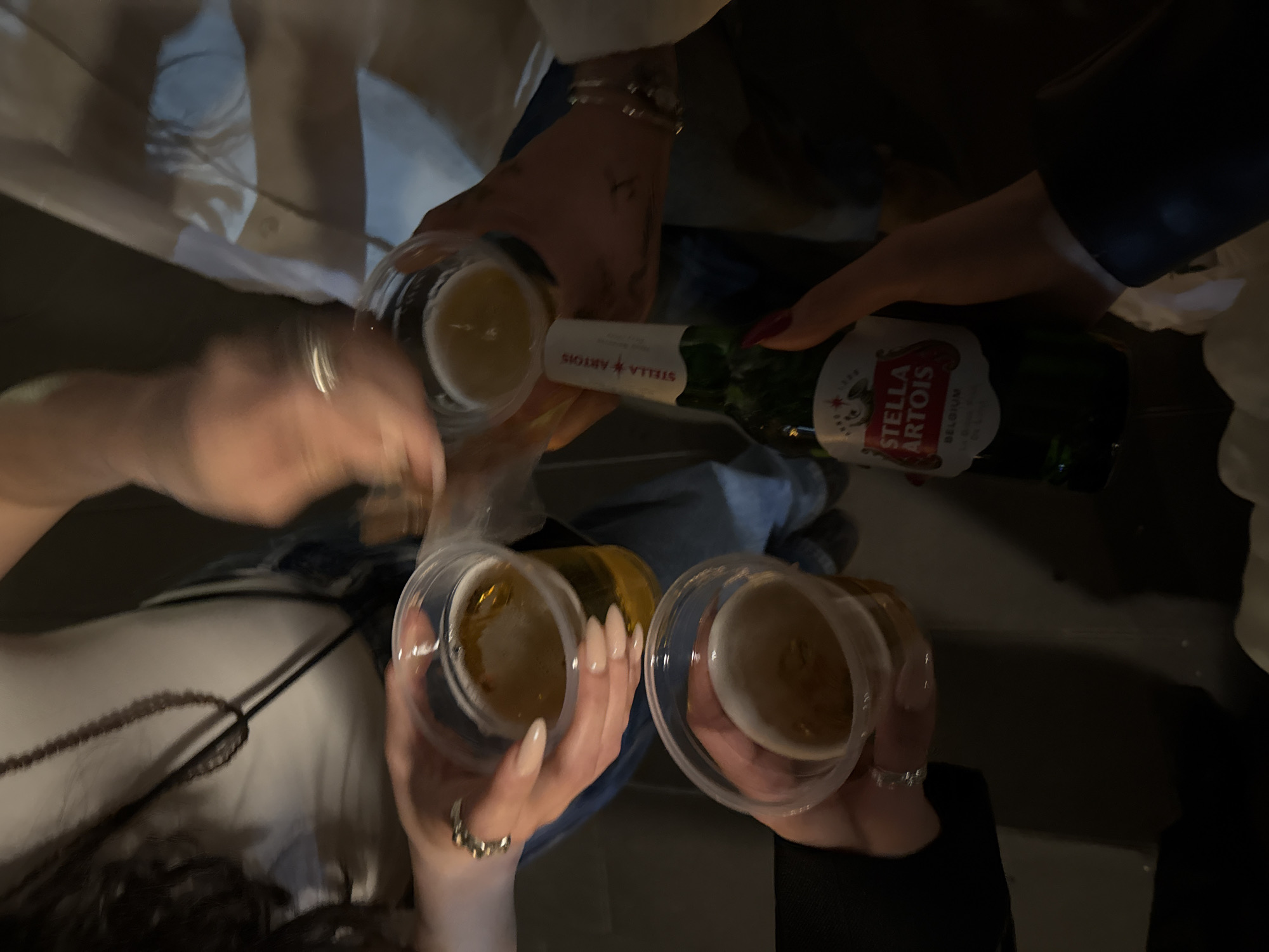 A slightly blurred overhead shot captures a group toasting with Stella Artois beer. The scene centers around a green bottle of Stella Artois being poured into three clear plastic cups, each filled with a yellowish liquid (presumably the beer). Hands holding the cups and the bottle are visible, some adorned with rings. Clothing styles suggest a casual setting. No information in the image or the hints allows for identification of Yerevan, Armenia.