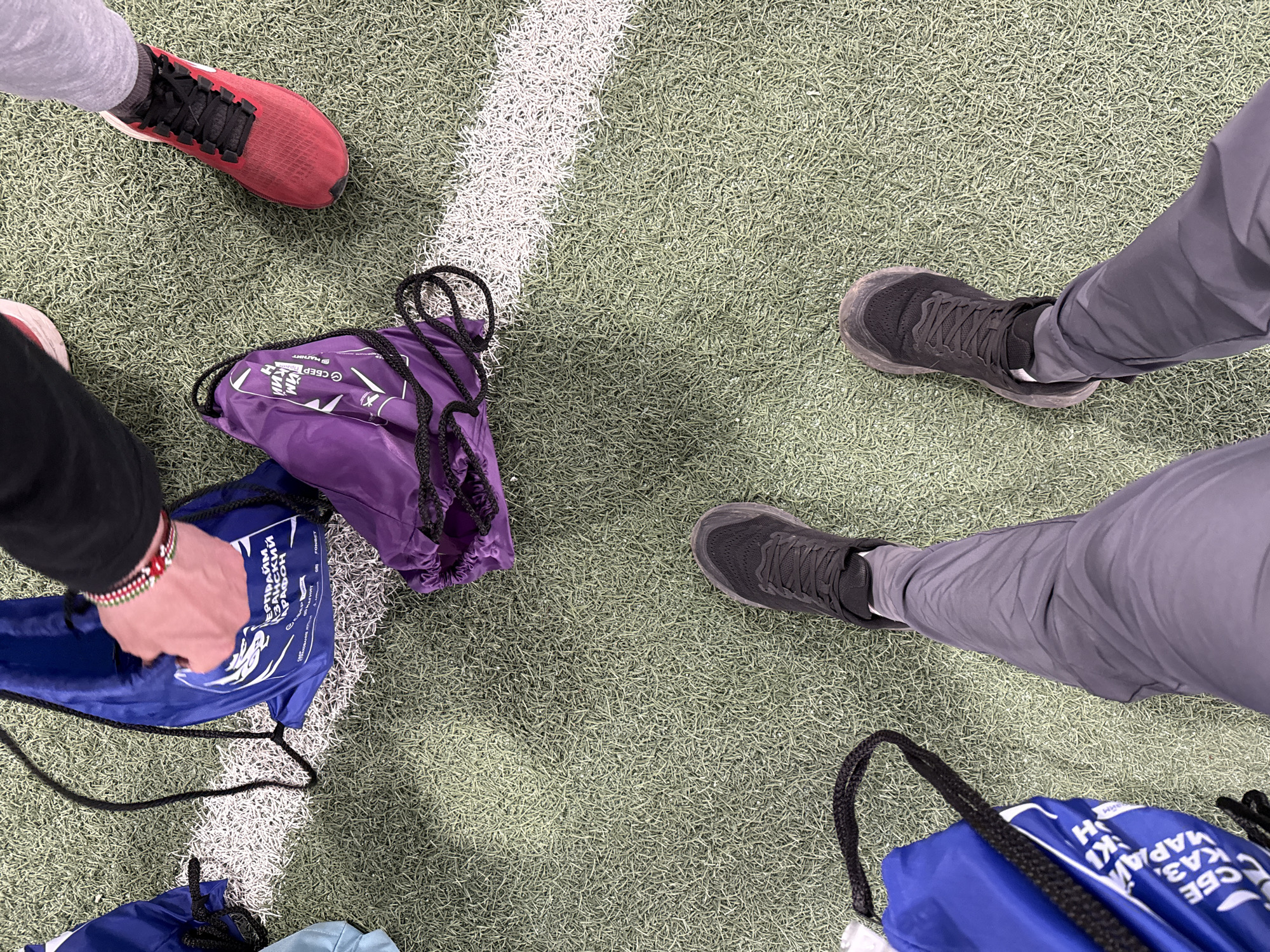 The image presents an overhead view of an artificial turf field. A white stripe is visible on the field. The scene includes sections of three individuals' lower limbs and footwear. One person is wearing red and black running shoes, another is dressed in gray pants and black sneakers, and the third person's arm and hand are visible. Several drawstring bags are strewn around the scene, colored purple and blue. The location in the city of Unknown, Unknown, is not defined by the image.