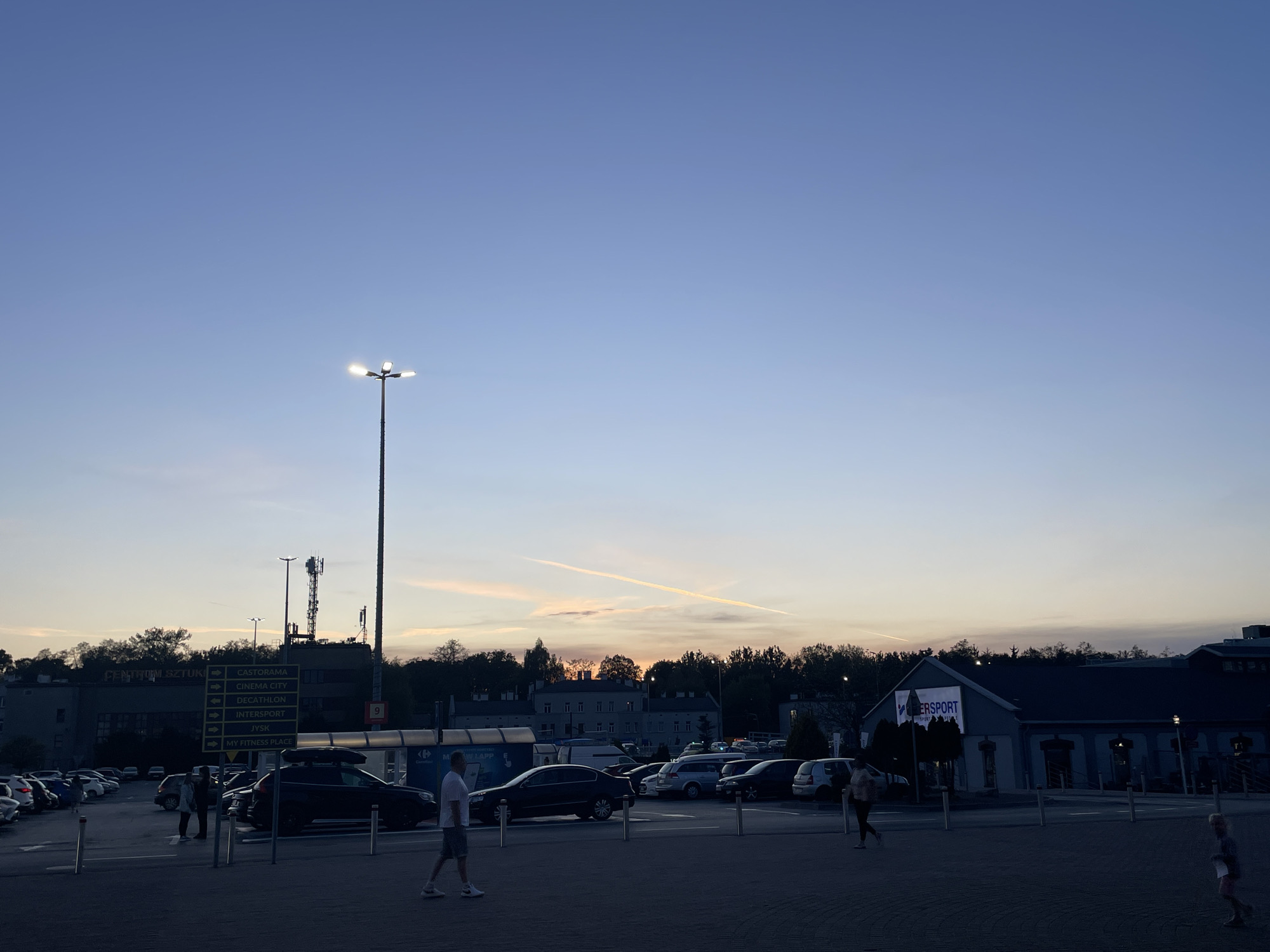 The image depicts an outdoor scene, likely a parking lot, at dusk. The sky is a gradient of blue, transitioning from a lighter hue near the horizon to a deeper blue overhead, with faint contrails of airplanes visible. Streetlights illuminate the foreground, casting a glow on the pavement. A tall pole with multiple lights stands prominently in the frame.
Several cars are parked in rows, and people are walking across the parking lot. A building with a sign that reads "INTERSPORT" is visible in the background, along with another building with a sign indicating directions to "CASTORAMA," "CINEMA CITY," "DECATHLON," "JYSK," and "MY FITNESS PLACE." The overall scene suggests a commercial area or shopping center parking lot at twilight. The location is Unknown, Unknown.