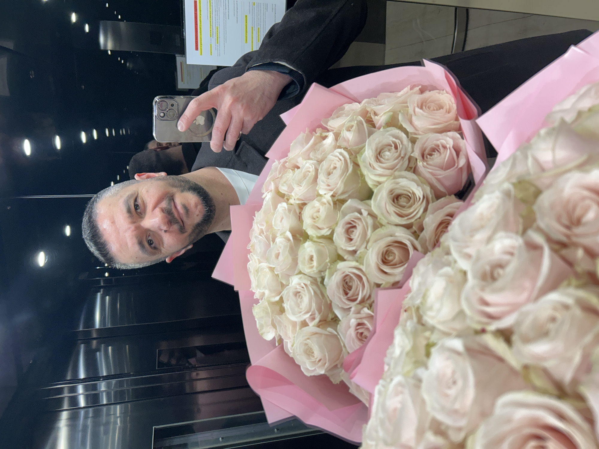 The image shows a man taking a selfie in what appears to be an elevator. He is holding two bouquets of pale pink roses wrapped in pink paper. The elevator has a black interior with horizontal metal accents and vertical lighting. A document is visible on the elevator wall. The location cannot be determined from the image alone.