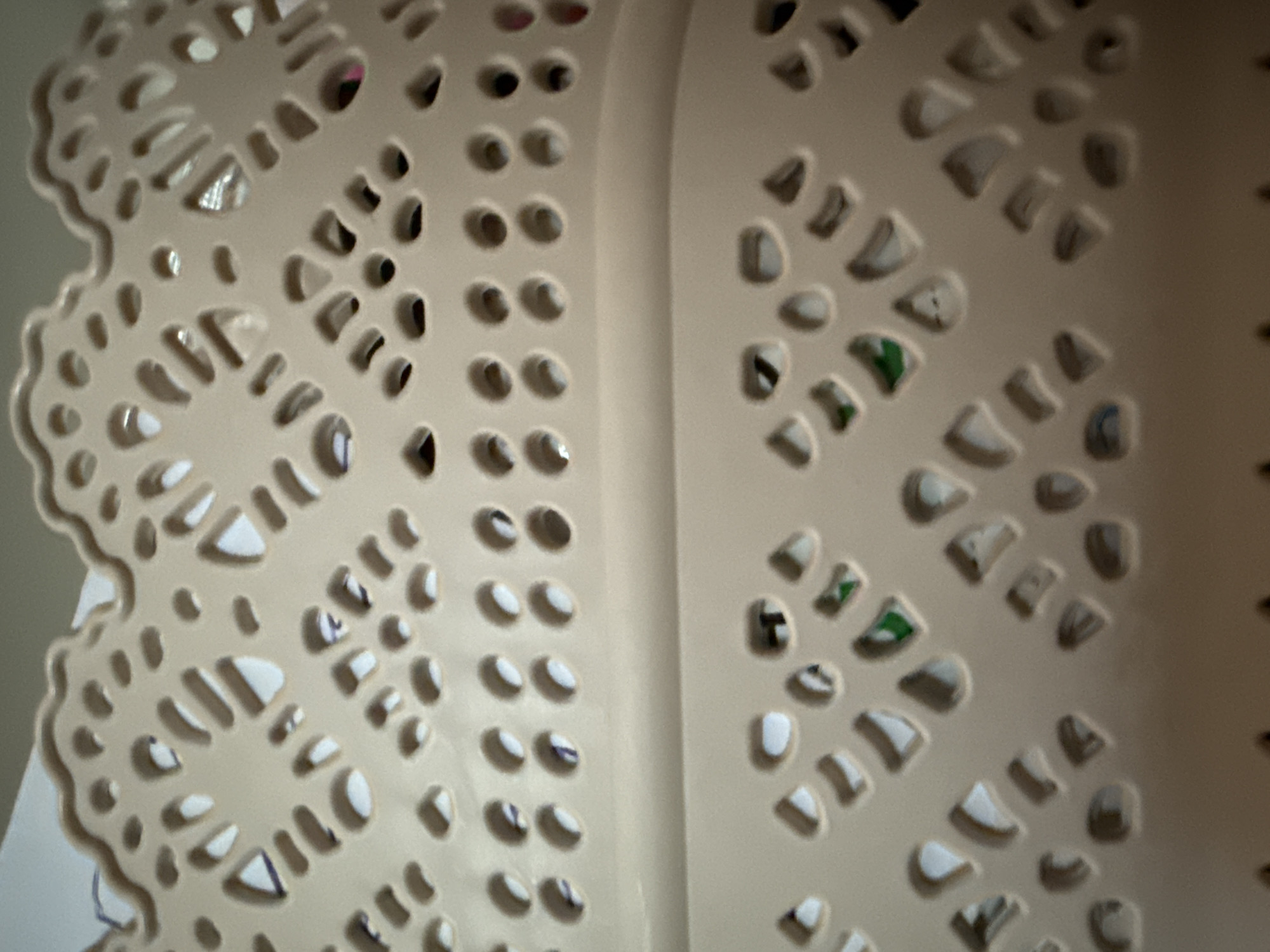 The image displays a close-up, slightly blurry view of a beige plastic basket with a decorative, openwork pattern. The basket appears to be made of a lightweight plastic and has a textured, possibly woven, design with many holes and cutouts. The top edge of the basket has a scalloped, lace-like finish. Through the holes, glimpses of various objects can be seen, including what appear to be pieces of paper, some with blue markings, and possibly some colored items like green paper or fabric. The lighting suggests an indoor setting, with some reflections visible on the plastic surface. The image does not contain any identifiable landmarks, people, or specific details that would allow for its location to be determined, including in Okhtyrka, Ukraine.