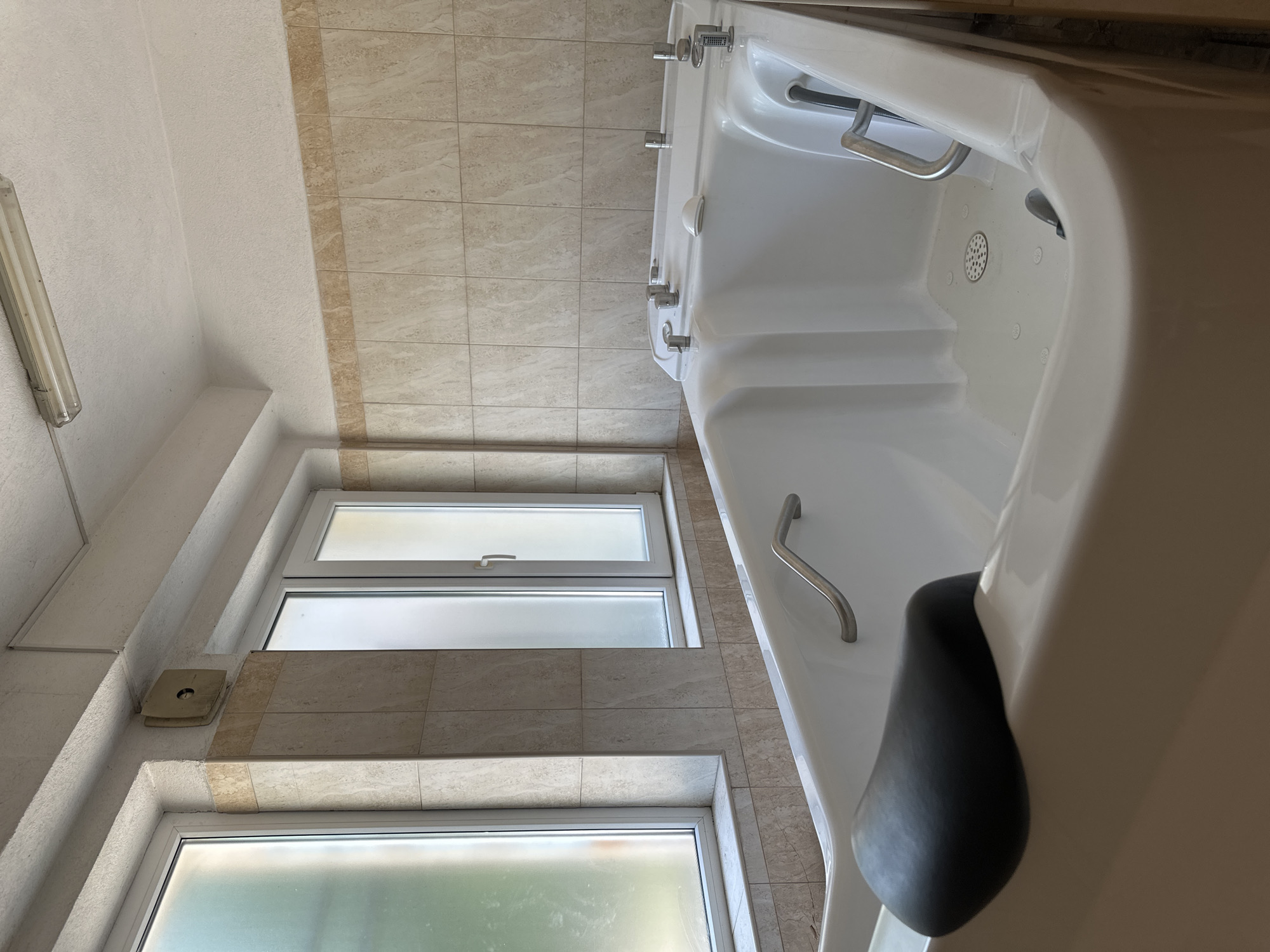 The image shows a bathroom setting with a white bathtub prominently featured on the right side of the frame. The bathtub has chrome handles and various controls along its edge. To the left of the tub, there are two windows with frosted glass. The wall behind the windows and the bathtub is tiled in a light tan color with a subtle pattern. A light fixture is visible on the ceiling in the upper-left corner. The ceiling and adjacent wall are painted white. A small, square vent is noticeable on the wall near the top window. Based on the architectural style and interior, the location is consistent with a bathroom in Sofia, Bulgaria.