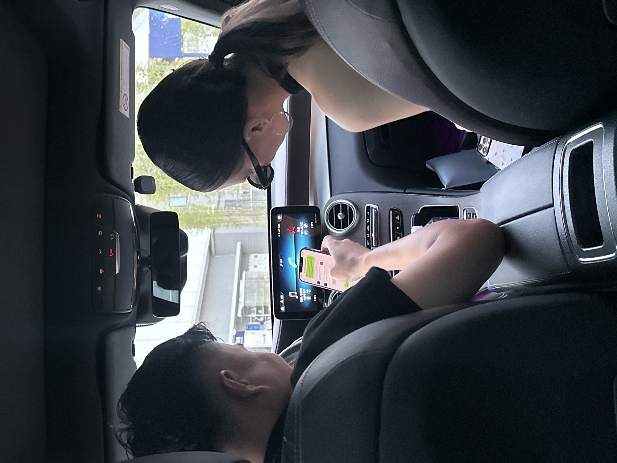 The image depicts two individuals inside a car, viewed from the back seats. In the foreground, a person with dark hair is holding a smartphone in their right hand, displaying a green-themed application, likely a messaging or social media platform. The phone screen is illuminated. The person is wearing a black t-shirt. In the background, another person with dark hair tied in a ponytail is wearing sunglasses and large hoop earrings, and a black top with a choker. The car's interior is visible, including the dashboard with a central infotainment screen displaying navigation or media information, air vents, and center console controls. The car is likely in motion or parked near a street, as indicated by the blurred outdoor scenery seen through the windows, featuring buildings and greenery. The presence of Chinese characters on the car's screen and the context provided suggests the location is in Liwan District, China.