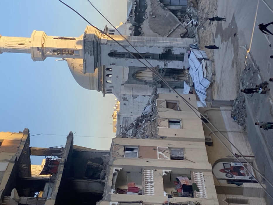 A minaret and domed building rise behind a partially destroyed building. Rubble litters the ground. The foreground structure has exposed rooms with hanging fabrics. A damaged advertisement is visible on its right side. Wires are strung across the sky. Small, dark figures walk along a road in the upper right, and other people mill on the ground nearby. The sky is a clear, light blue. The light suggests a late afternoon or early morning scene.
