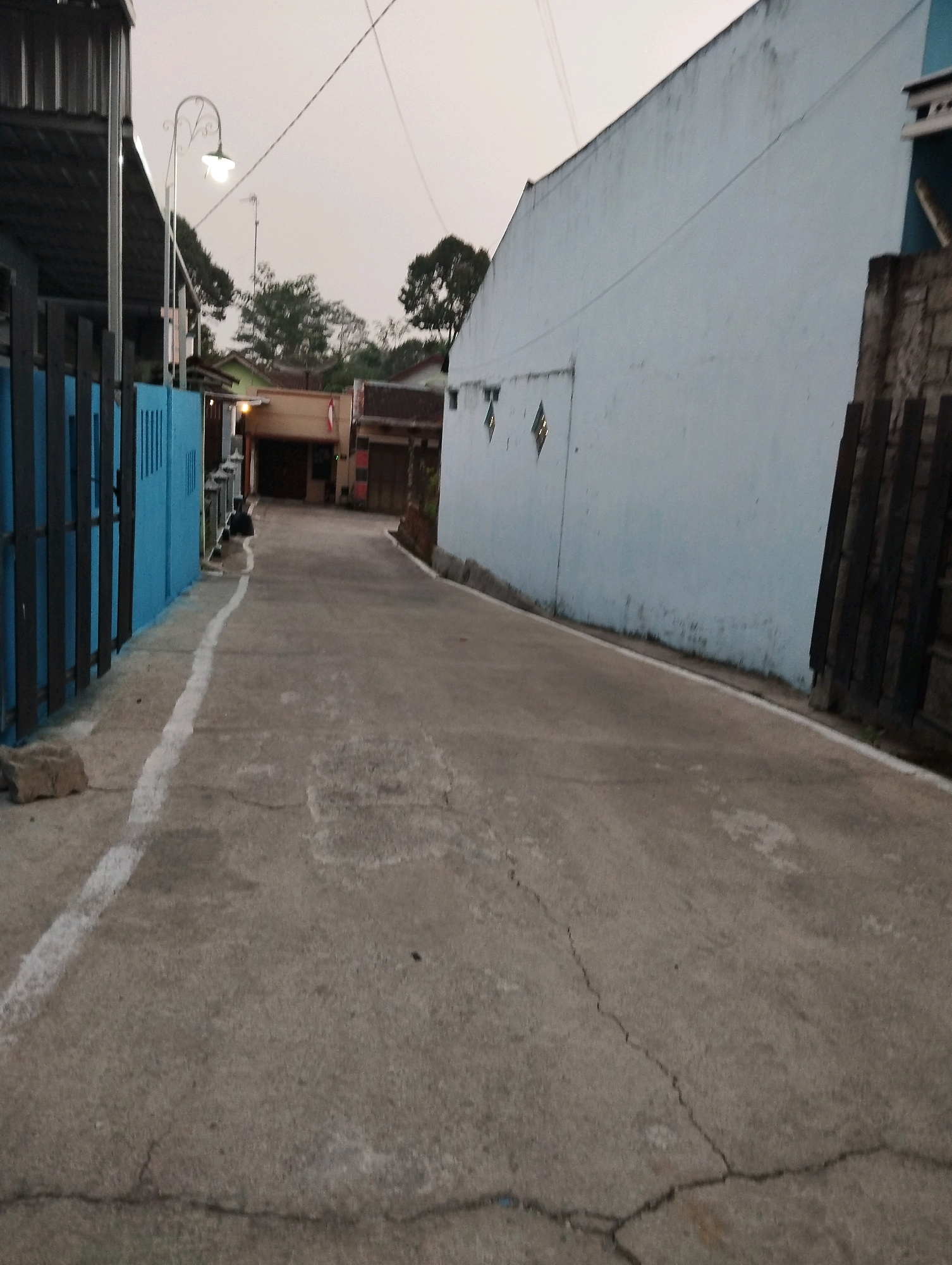 The image depicts a narrow paved alleyway in Salatiga, Indonesia, with buildings flanking both sides. On the left, a bright blue fence borders the alley, behind which is a structure with a metal roof. A street lamp with a decorative curved arm is visible above the fence. The right side of the alley is dominated by a large, light blue wall that extends into the background. A section of dark wooden fence or wall is situated in the foreground on the right. The alleyway itself is made of concrete, showing cracks and wear, with a white painted line marking the edge on the left side. In the distance, more buildings with tiled roofs and glimpses of trees are visible under a hazy, overcast sky, suggesting it is either early morning or late afternoon. The scene is quiet and appears to be a residential or commercial area.